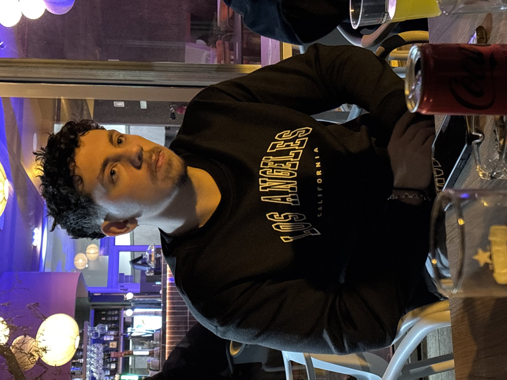 A man with curly dark hair, a light complexion, and stubble is seated at a table, possibly in a bar or restaurant. He is wearing a black sweatshirt with "Los Angeles California" written on it in stylized white lettering. He looks towards the camera. A dark-colored can and several glasses are on the table in front of him. In the background, there is a bar area with shelves of liquor bottles and decorative lighting. The location is possibly in Vila Real, Portugal, though there is no immediately identifying information in the image that confirms this.