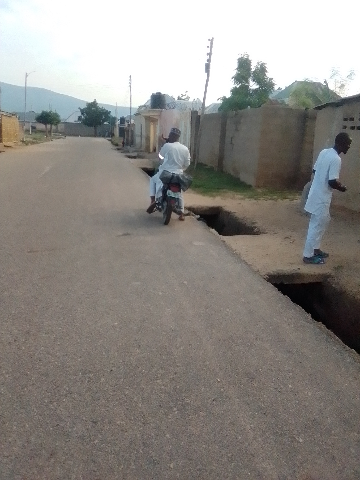 The image depicts a scene in Liji Hill, Nigeria, with two individuals present. The first individual is riding a motorcycle down a paved road, with another person seated behind them. Both are wearing white garments. The motorcycle appears to have some luggage secured to the rear. To the right of the road, a second individual, also wearing white, stands on the edge of a significant trench or erosion feature that runs alongside the road. This trench is deep and has steep, uneven sides, indicating potential danger. The environment is characterized by a road, adjacent buildings constructed from what appears to be mud brick or similar materials, and some vegetation, including trees in the background. The sky is overcast, suggesting it might be early morning or late afternoon. The overall impression is a street in a residential or semi-rural area, showing signs of natural erosion impacting the infrastructure.