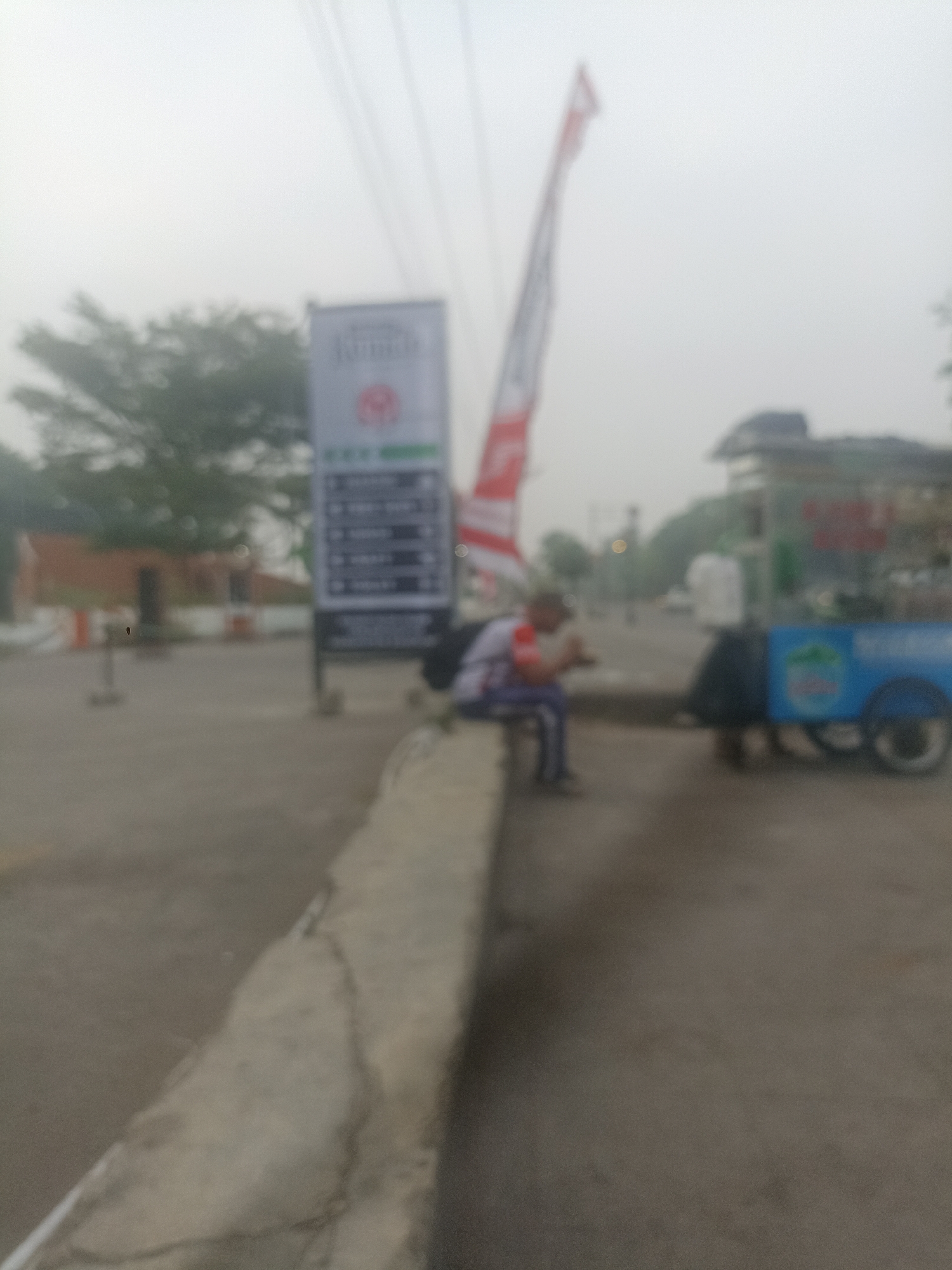 The image depicts an outdoor scene with a person seated on a concrete barrier. The person is wearing a white shirt with red accents and blue pants. They are looking down, possibly at a mobile device. To the right of the seated person is a blue cart with a white canopy, resembling a food or drink vendor. In the background, there's a tall sign with text and a circular logo, and a red and white flag is visible to the left of the sign. Trees and buildings are discernible in the distance. The overall environment appears to be a public area, possibly a roadside or a town square, with paved ground. The image is slightly blurred, suggesting it may have been taken in low light conditions or with a moving camera. The location is identified as Pamekaran, Indonesia, which suggests this is an urban or suburban setting.