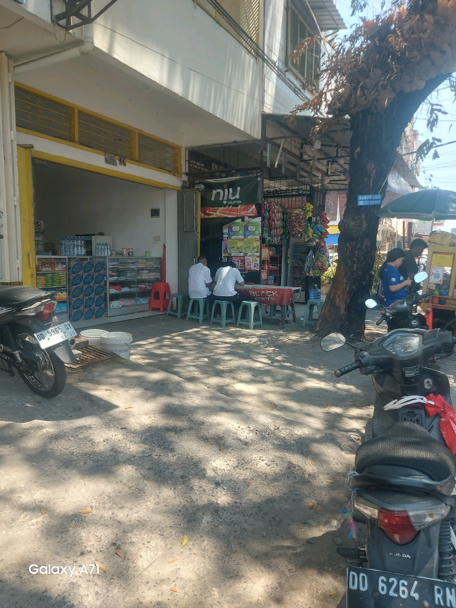 This is a medium shot of a street scene in Makassar, Indonesia, on a sunny day. The foreground is dominated by two parked motorcycles, a dark-colored one on the left and a newer model on the right. The motorcycle on the right has a black mesh seat cover and a red fabric tied to the back. A white bag with red trim is also visible. In the background, there is a small shop with an open front, displaying various goods on shelves. Two men are seated at a low table in front of the shop, appearing to be eating or conversing. One is wearing a white shirt, and the other a patterned shirt. Further back, a woman in a blue shirt is interacting with someone near a food cart. A large tree with dry leaves stands to the right of the shop. The ground is paved with asphalt and dirt, with shadows from the tree and structures cast across it. A sign on the tree reads "SUNUR BOR 085 289 499 000". The overall impression is one of a bustling, everyday street scene in a commercial area.