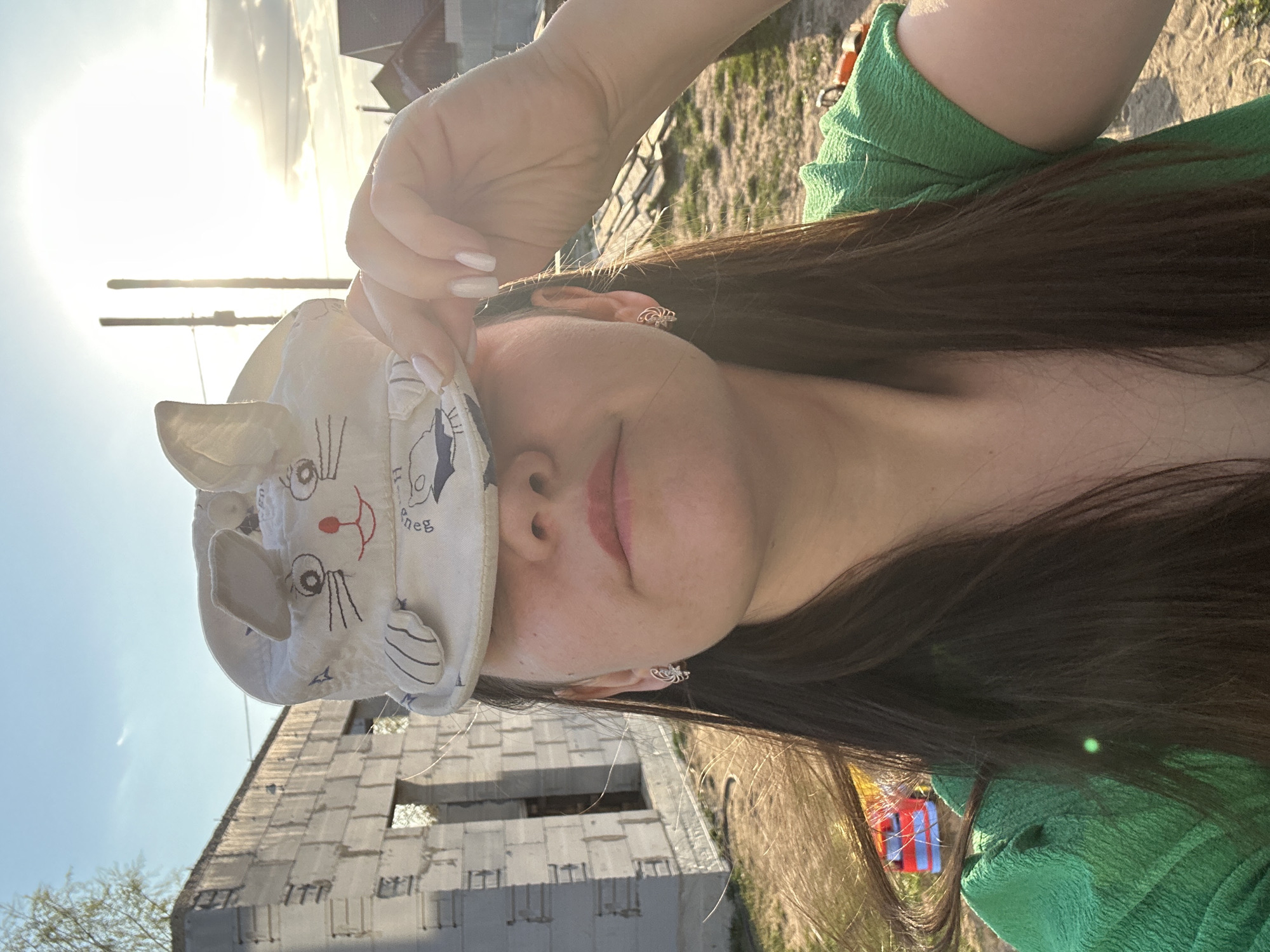 A woman with long, dark hair wearing a green textured top is holding a white cap with a cartoon bunny face design over the upper half of her face. She is outdoors on what appears to be a sunny day. In the background is a building under construction made of light-colored blocks, and further back is grassy terrain. Based on the location details provided, this scene is in Chaikovsky, Russia.