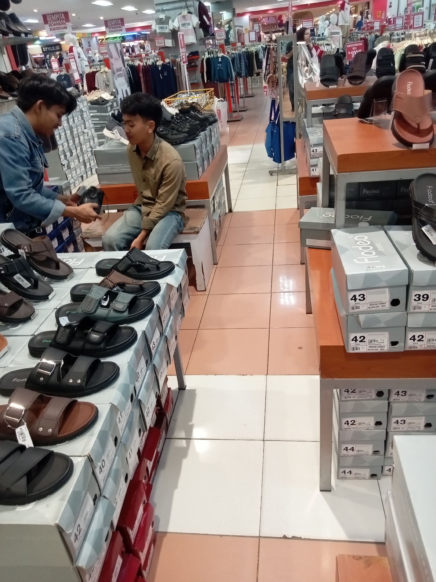 The image shows a retail shoe store in Batam, Indonesia, likely at a mall like Nagoya Hill or Grand Batam Mall, judging by the store's interior design and display styles common in Indonesian department stores. Two young men are the main subjects in the foreground. The man on the left, wearing a denim jacket and blue jeans, is holding a black shoe in his hands, appearing to show it to the man on the right. The man on the right, dressed in a brown button-down shirt and blue jeans, is sitting on a low bench and looking at the shoe. They are in front of a display of various sandals and shoes, many of which are arranged on top of shoe boxes. The shoe boxes are stacked neatly, with some displaying shoe sizes like '43', '42', and '40'. The store environment is bright, with overhead fluorescent lighting illuminating the aisles and product displays. Clothing racks with apparel can be seen in the background. The floor is tiled, a common feature in commercial spaces. Several pairs of sandals are displayed on shelves, in various colors and styles, including flip-flops and more structured sandals with straps. A blue shopping bag is visible in the background, indicating active shopping. The overall scene captures a moment of footwear selection and potential purchase in a bustling Indonesian retail setting.