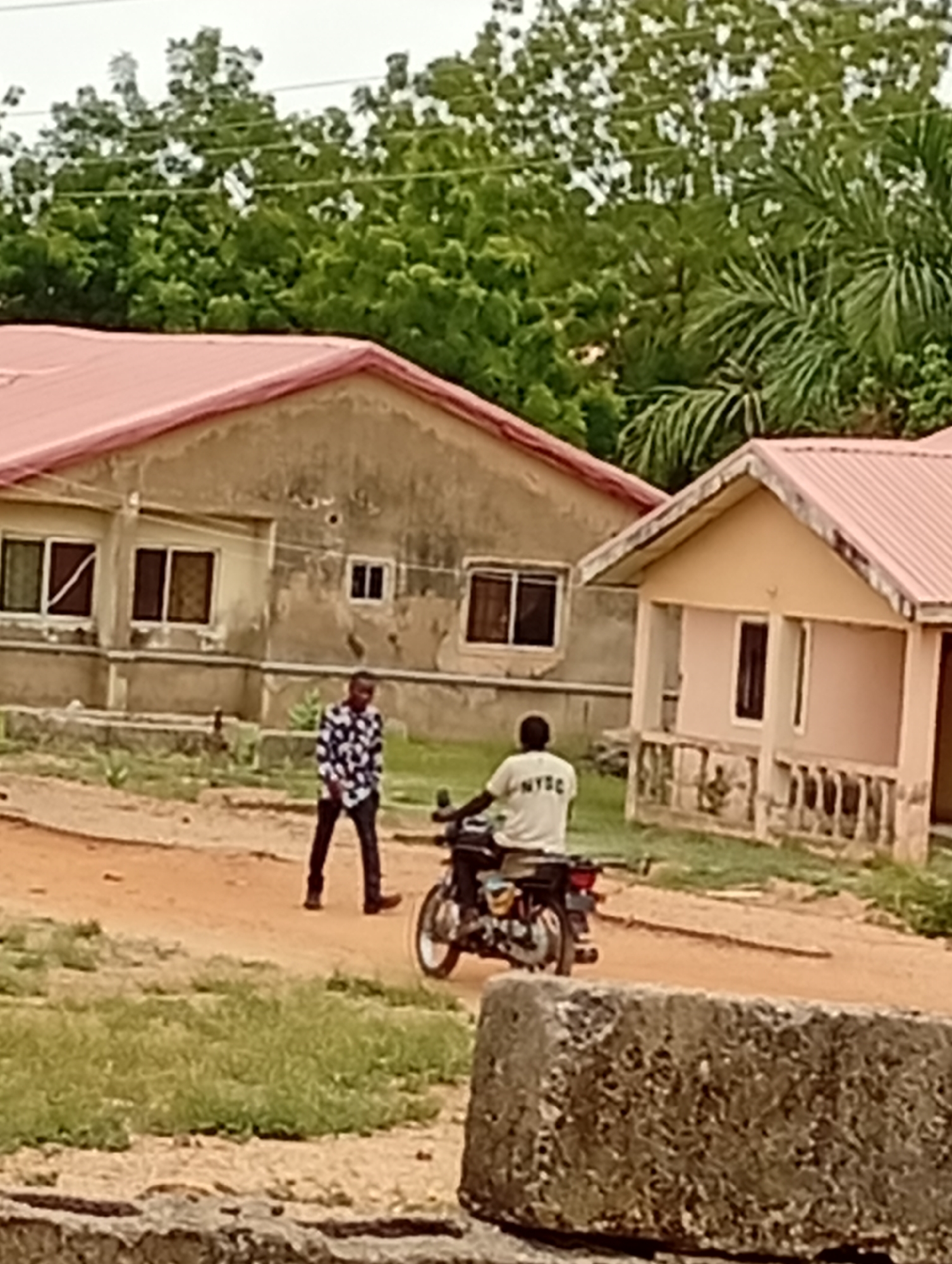 The image depicts a street scene in Adankolo, Nigeria. A man wearing a black and white patterned shirt and black pants walks on a dirt road. A person on a motorcycle, wearing a white shirt with "NYSC" printed on the back, is also on the road. In the background, there are two houses with red roofs. The first house has a weathered exterior with visible windows. The second house is smaller, with a porch area supported by pillars. Green trees and foliage are visible behind the houses. In the foreground, a weathered stone block partially obscures the view.