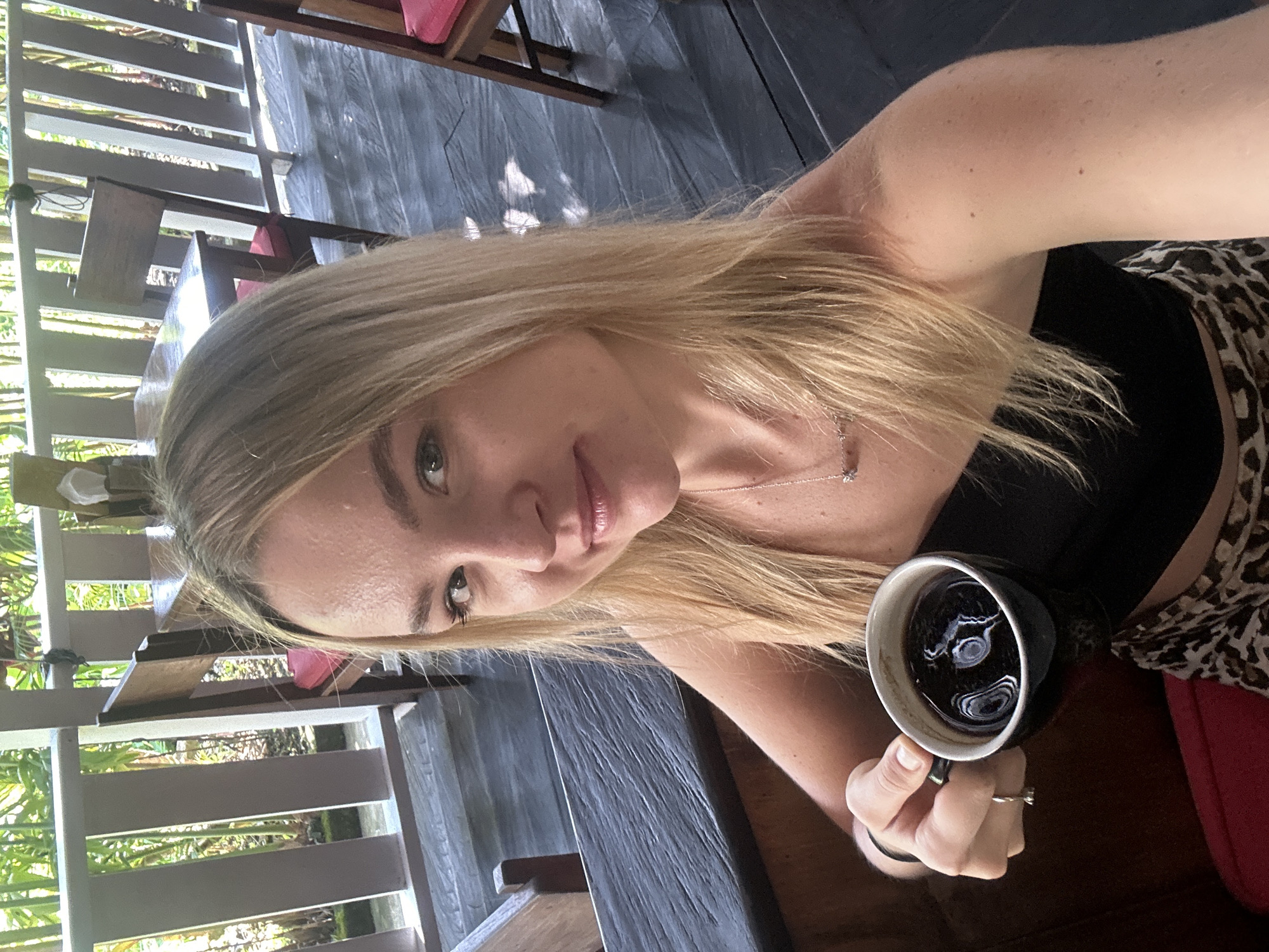 A blonde woman is taking a selfie while holding a cup of coffee. The setting appears to be an outdoor cafe in North Kuta, Indonesia. The woman is wearing a black top and a leopard-print skirt or shorts. She is seated at a wooden table on a wooden deck with railings. Tropical foliage is visible beyond the railings.