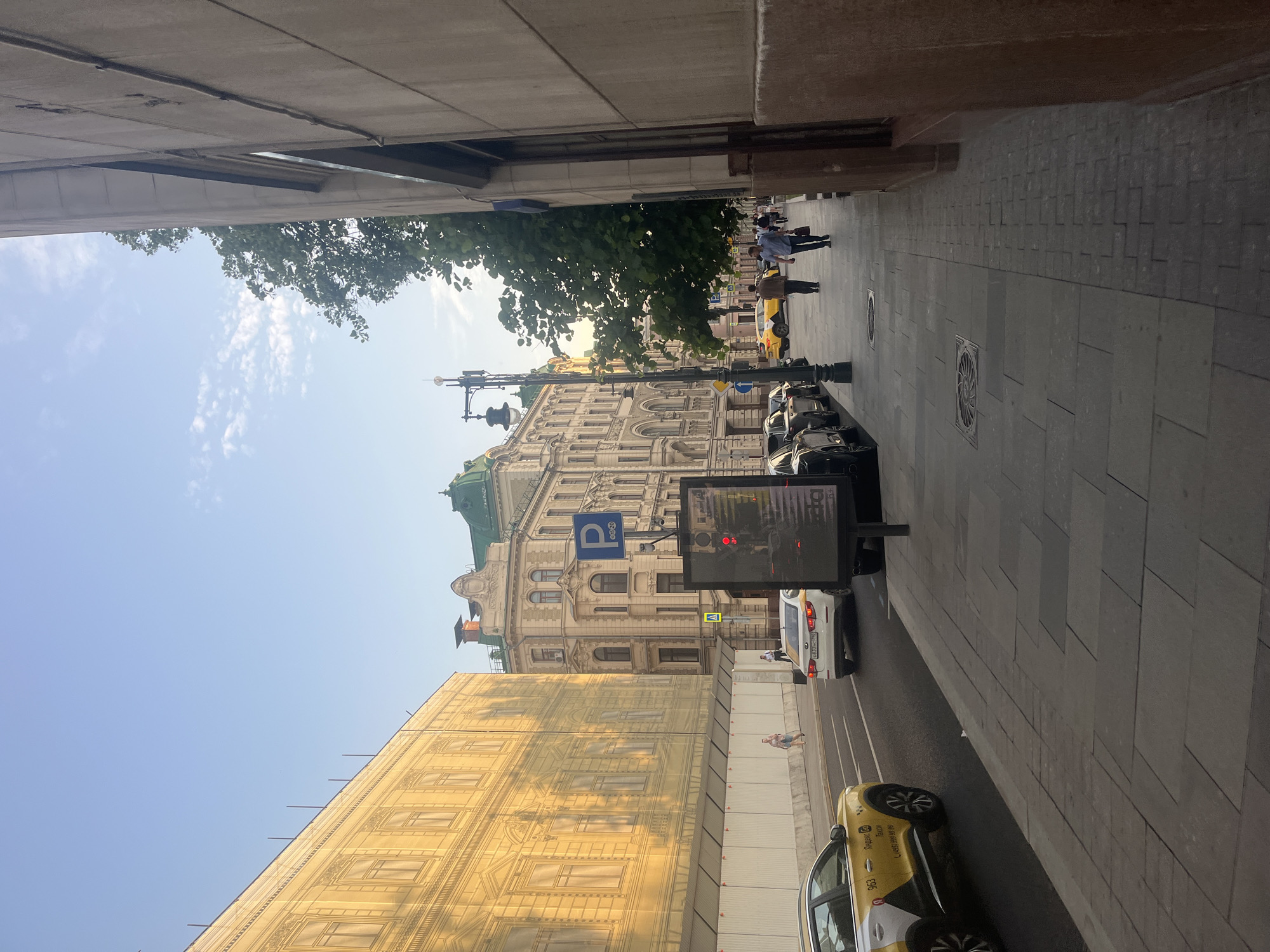 This image captures a street scene in Moscow, Russia. On the left, a large building is partially covered by a yellow tarp that mimics its architectural facade, with several windows visible. Adjacent to it is another building with intricate architectural details and a green roof. In the foreground, a sidewalk runs along the street. Several cars are present, including yellow taxis with Russian markings, and other passenger vehicles. Pedestrians are walking on the sidewalk. A tall lamppost with a decorative fixture stands near the sidewalk. A digital billboard displays a red traffic light. The sky is clear with a few wispy clouds. The overall impression is a bustling city street during daylight.