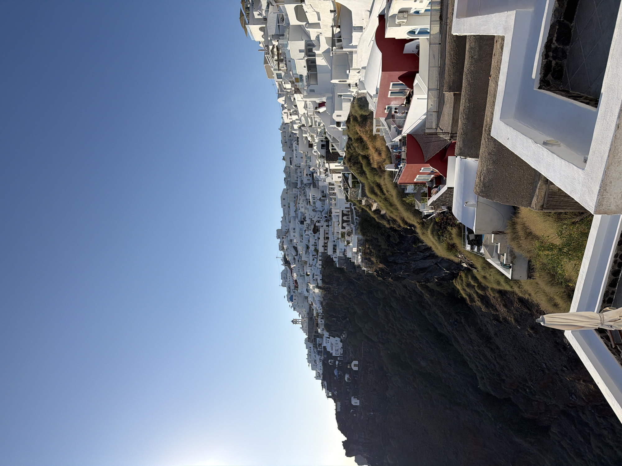 The image shows a panoramic view of Oia, a village in the Santorini island in Greece. The village is situated on a cliff overlooking the Aegean Sea. The houses are whitewashed and built in a traditional style, with blue domes and narrow streets. The sky is clear and blue, and the sun is shining. The cliff face is covered in vegetation. In the foreground, there are some white buildings and a white umbrella. In the middle ground, there are more white buildings, some with red accents, and some people on a balcony. In the background, the village continues up the cliff face.