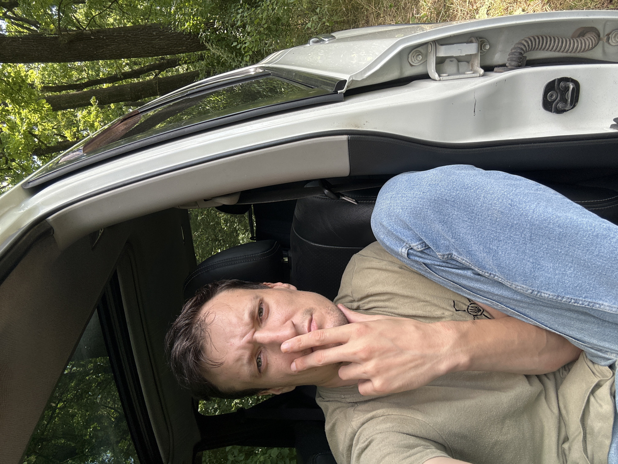 A man sits in the driver's seat of a white SUV, partially obscured by the open passenger door. He wears a tan t-shirt and blue jeans, with his left arm resting on his lap and his right hand held up to his face, obscuring his nose and mouth. The vehicle is parked outdoors, with lush green trees and foliage visible through the sunroof and the open door, suggesting a wooded or rural setting. The man's facial expression is one of apparent frustration or contemplation. The scene appears to be in Makiv, Ukraine, based on the context provided.