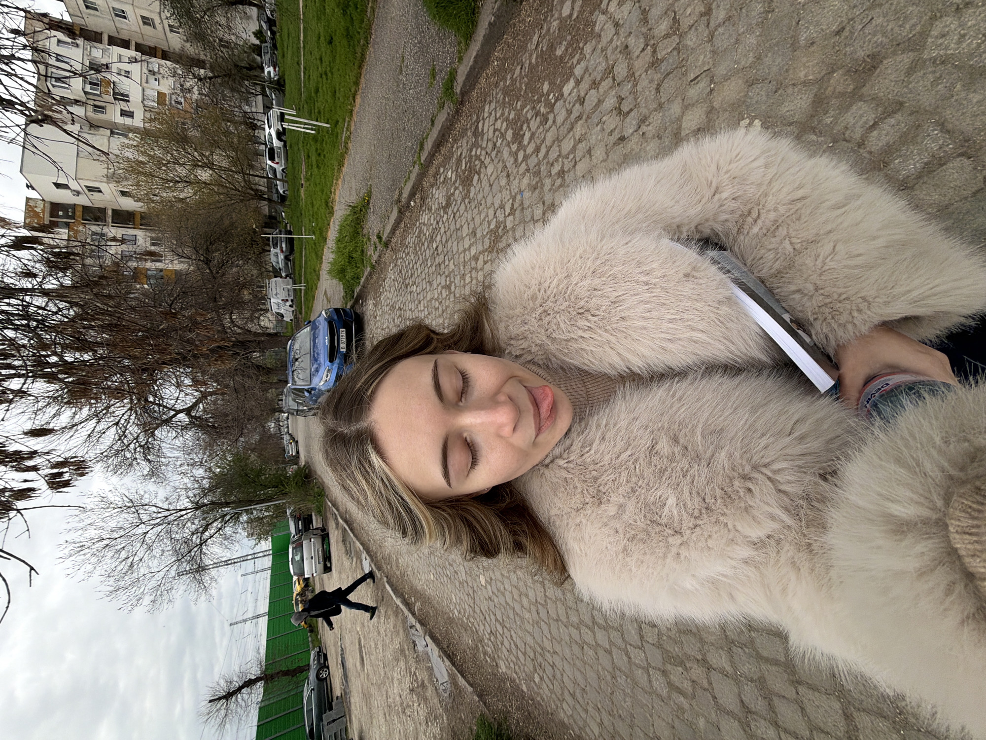 A young woman lies on a cobblestone path in Plovdiv, Bulgaria. She is wearing a light beige fur coat and holding a book or magazine. Her eyes are closed, and she has her tongue slightly out. In the background, there is a street with parked cars and buildings. A person is walking in the background.