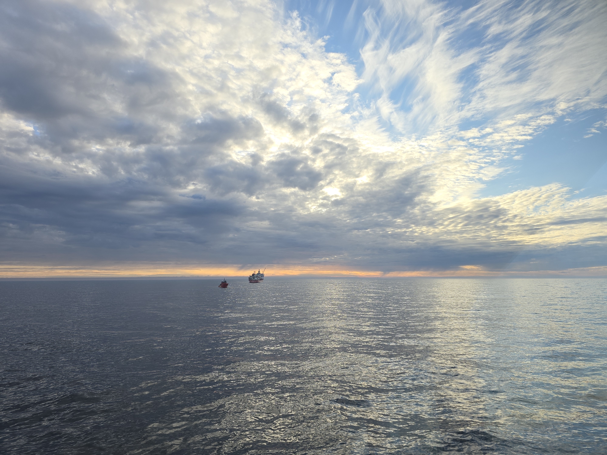 The image depicts a seascape under a sky with a mix of dark, low-lying clouds and patches of blue sky streaked with wispy clouds. The sun appears to be setting or rising along the horizon, casting a warm, orange glow. Two vessels are visible on the water. One is a small, red boat, and the other is a larger ship or platform in the distance. The water itself is calm, with gentle ripples across the surface.