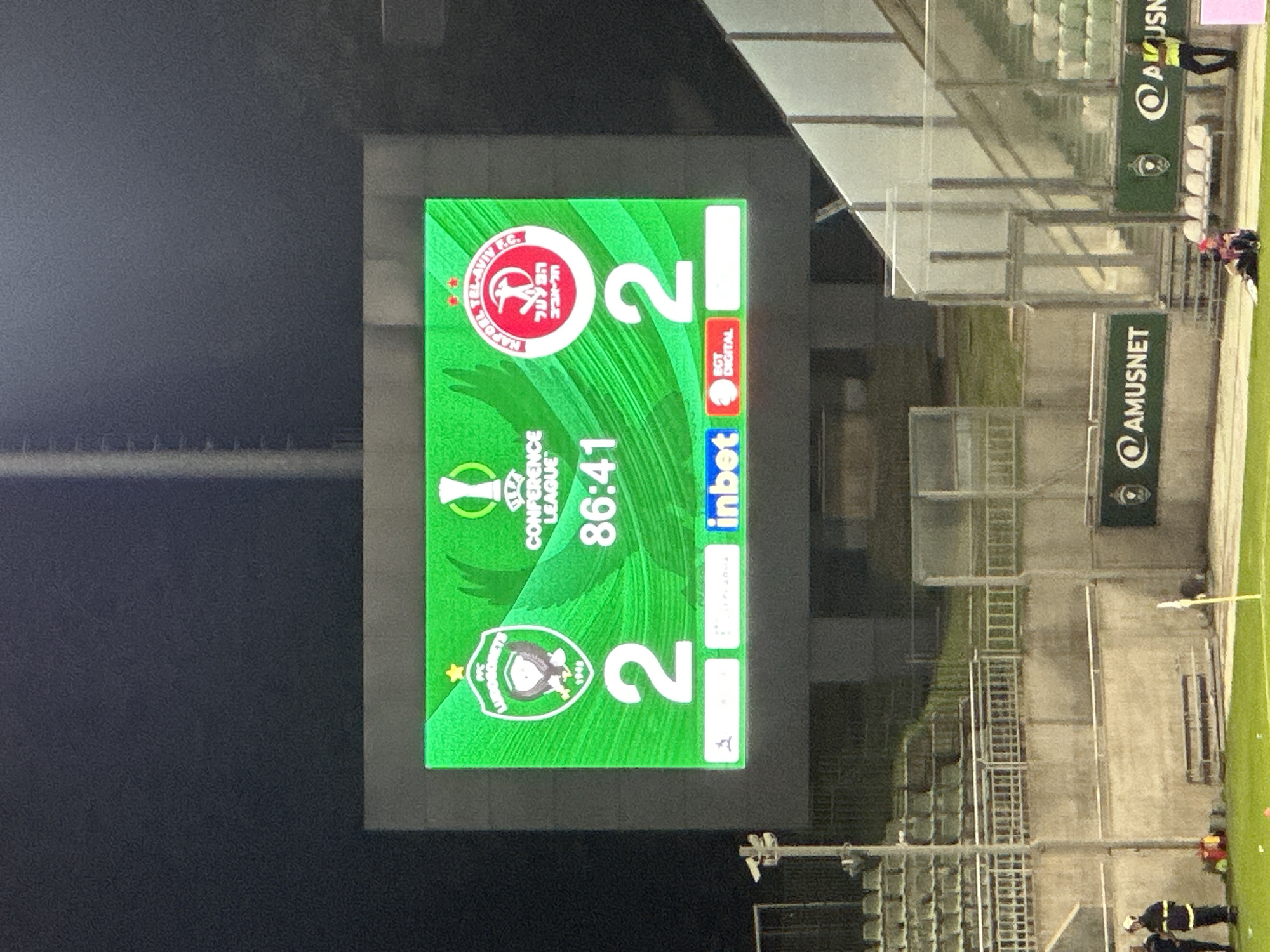 The image shows a scoreboard at a football stadium, likely in Razgrad, Bulgaria. The scoreboard displays a match between PFC Ludogorets and Hapoel Tel Aviv F.C. The score is 2-2 at 86 minutes and 41 seconds. The logos of the Conference League, PFC Ludogorets, and Hapoel Tel Aviv F.C. are visible. Below the main score, there are sponsor logos for "inbet" and "BGT DIGITAL". The stadium seating is visible in the background, as are some security personnel and a few spectators in the foreground. The overall environment is an outdoor football stadium at dusk or night, with artificial lighting illuminating the field and scoreboard.