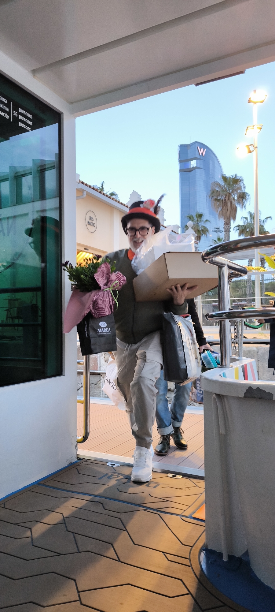 A man in a dark green jacket, light grey pants, and white sneakers is carrying a large brown box, a bouquet of flowers in a pink wrapping, and multiple shopping bags, including one from Marea, while stepping through a doorway. He is also wearing a hat with feathers and glasses. Behind him, another person is partially visible, wearing jeans and boots. The setting appears to be a boat or dock, with the floor having a patterned wooden design. In the background, there is a building with a "W" logo on it, suggesting the W Barcelona hotel. The building and palm trees point to the city of Barcelona, Spain. The lighting suggests it is late afternoon or early evening. There are railings and boat fixtures visible, and a sign indicating the boat's capacity as 56 persons.
