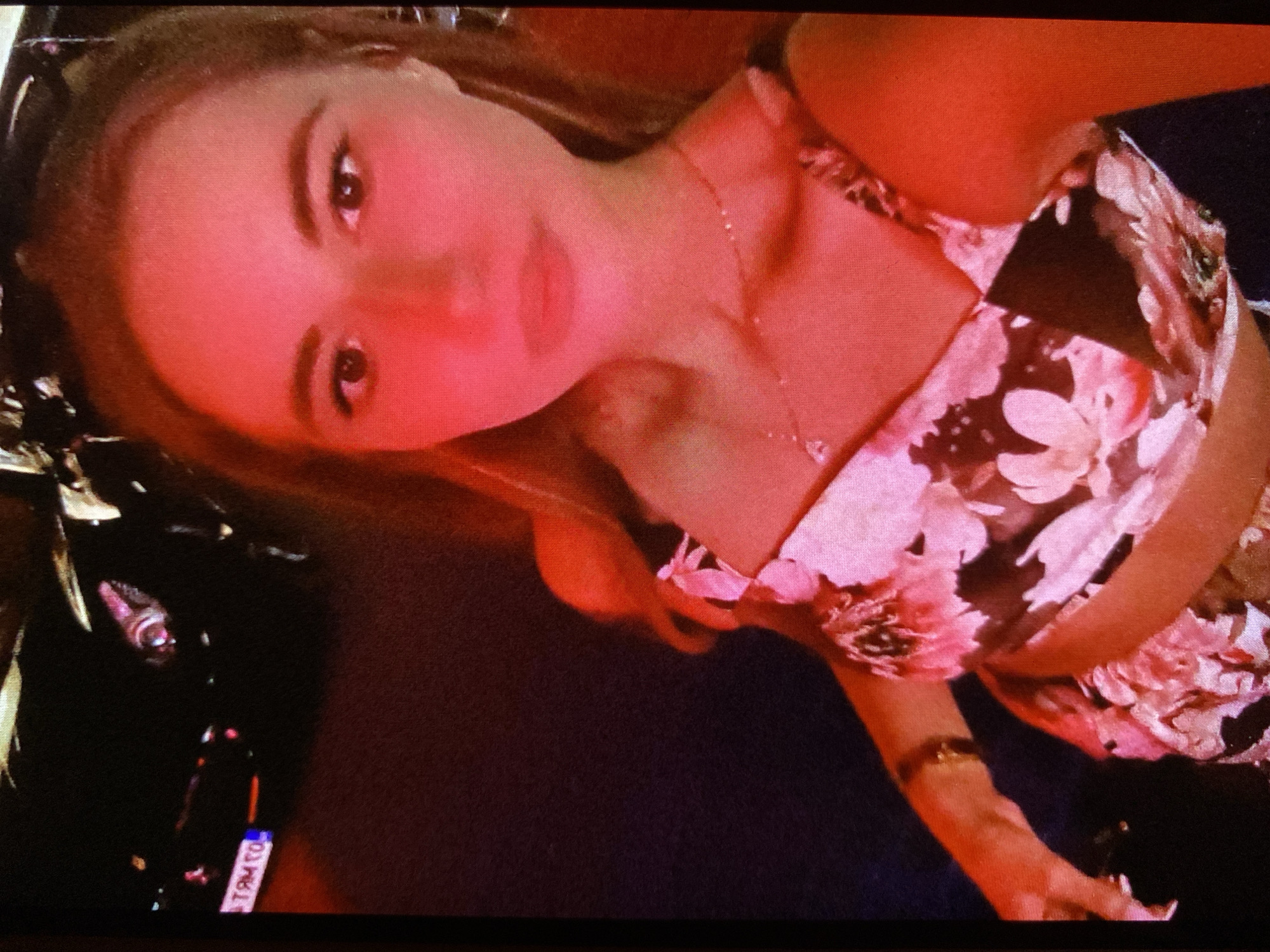 The image displays a young woman posing for a photo, seen at an angle due to the tilted orientation of the shot. Her face is illuminated by a dominant reddish-pink light. She has light brown hair, dark eyes, and well-defined eyebrows, looking directly at the viewer with a neutral to slightly smiling expression.
She is wearing a sleeveless crop top or bralette featuring a floral pattern in shades of black, pink, and white. A delicate gold-colored chain necklace with a small pendant is visible around her neck, and a matching bracelet adorns her left wrist. Her fingernails appear to have a light polish. Sunglasses are pushed back on her head, partially visible on the upper left side of the frame.
The scene is dimly lit, suggesting a night-time environment, possibly indoors or at an outdoor event in Bankya, Bulgaria, given the location context. The background is dark and indistinct, with some metallic or reflective elements visible on the far left. The overall lighting scheme indicates artificial, colored illumination, likely from a stage light or decorative lighting.
In the bottom left corner, a small, white rectangular tag or label is visible. The text on it is partially obscured and mirrored, but can be deciphered as "OT MA9 TO," potentially "TO 9AM TO" when correctly oriented. This could be an event tag, a label on an object, or similar informational text. The atmosphere suggests a relaxed, social setting.