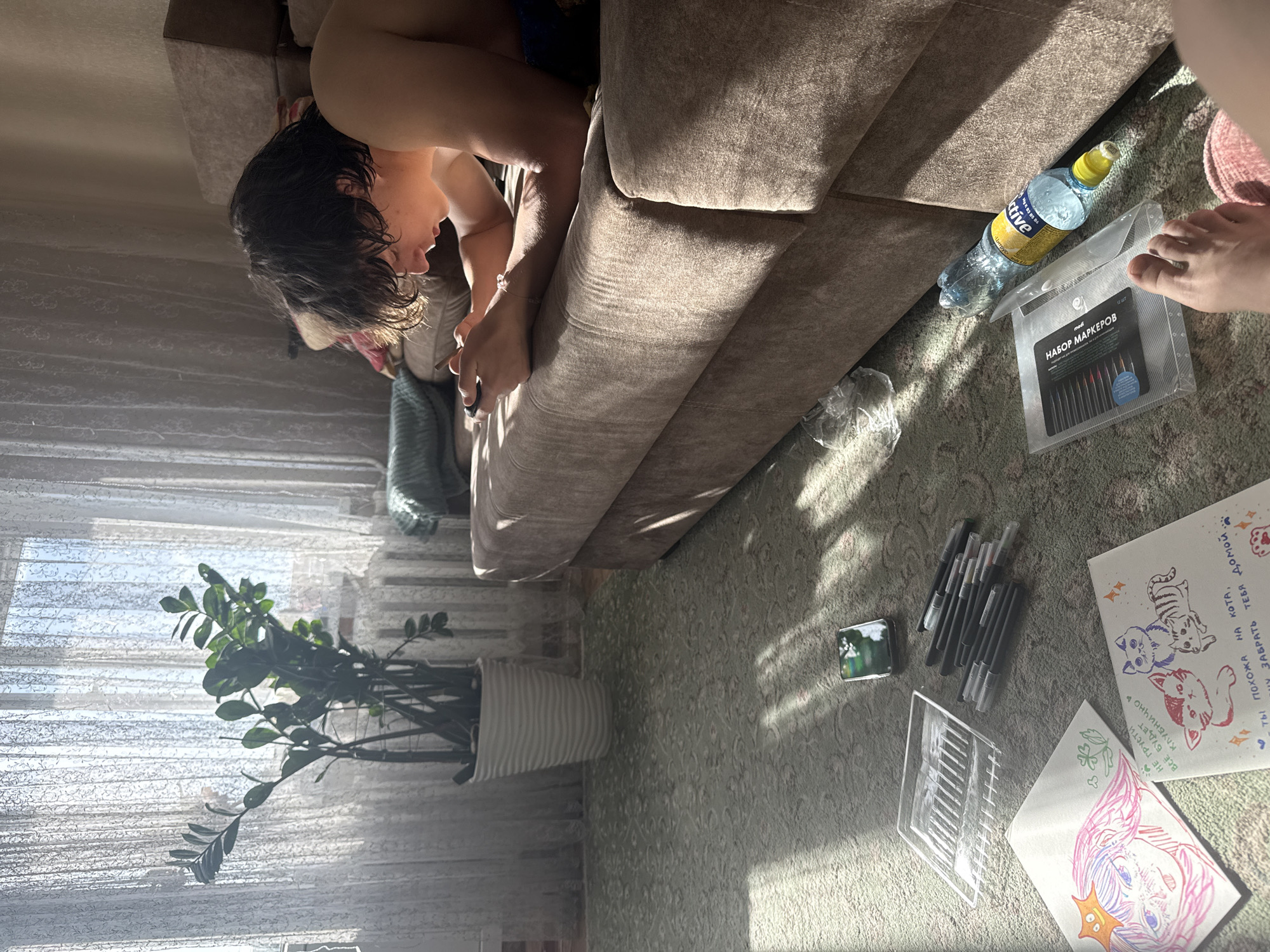 The image shows a person sitting on a brown sofa, possibly drawing or using a mobile phone. Next to the sofa on a carpeted floor, there is a bottle of water, a set of markers, several loose pens, and two drawings. One drawing features three cats in different colors, and the other depicts a character with pink and purple hair. A potted plant is visible near a window with sheer curtains. The texts on the drawing are written in Russian. Based on the marker brand medi and the Russian text, it's likely the location is in Russia. Given that some of the text mentions "strawberry" and given that Урюпинск (Uryupinsk) in Russia is known for producing strawberries, it is likely the location is in Uryupinsk, Russia.