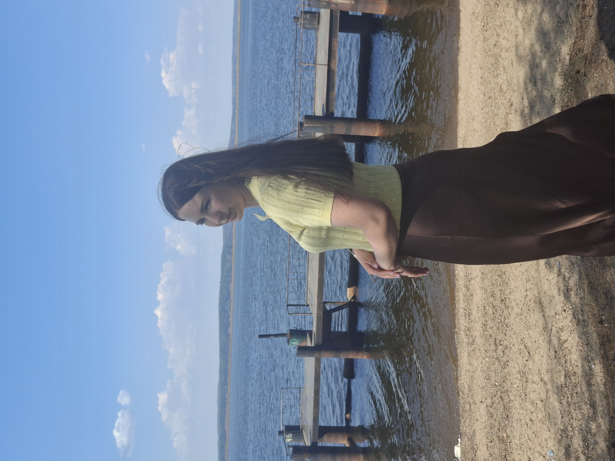 A woman with long brown hair is standing on a sandy beach next to a body of water under a blue sky with a few white clouds. She is wearing a light green shirt and a brown skirt. Her hands are clasped in front of her. In the background, there is a pier or a structure extending into the water, supported by metal posts. The location is Chaikovsky, Russia.