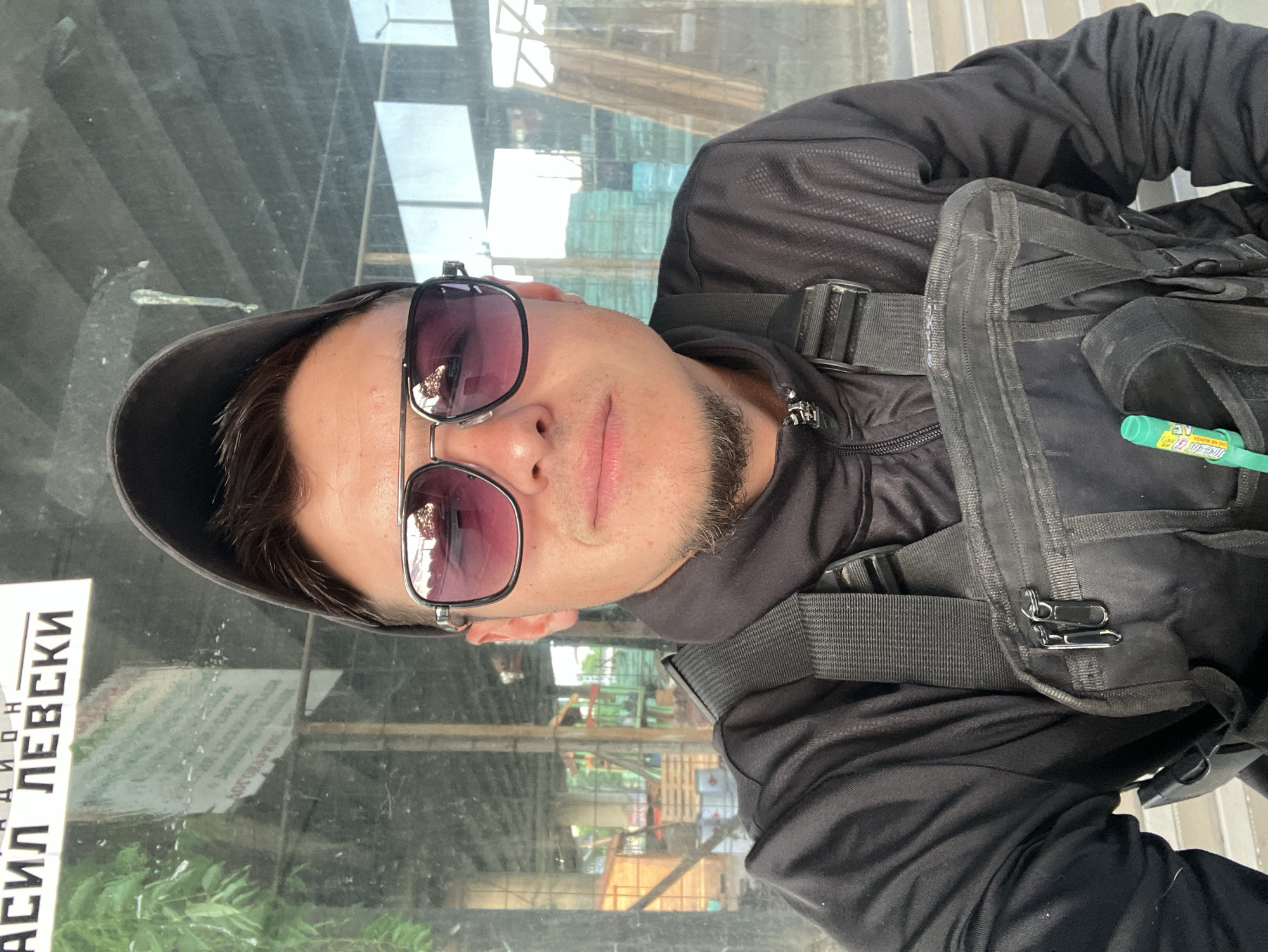 A medium shot captures a man wearing sunglasses and a black cap and jacket, with a black bag or vest over his upper body. The reflection in the glasses suggests an outdoor setting, possibly an urban area. The man appears to be standing or sitting in front of a glass partition. The reflection in the glass reveals a building or structure with scaffolding. The location is Plovdiv, Bulgaria, as evidenced by the Cyrillic text "АСИЛ ЛЕВСКИ" visible in the reflection, indicating the name of a street or place in Bulgarian.