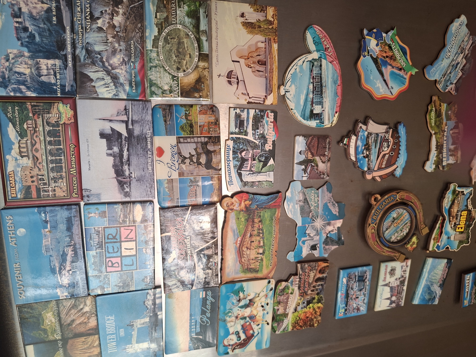 The image displays a collection of refrigerator magnets, primarily featuring landscapes, architecture, and some illustrations. The magnets are arranged on a dark, metallic surface, likely a refrigerator door.
The subjects include diverse scenes such as natural landscapes with mountains and water bodies, historical buildings like monasteries and churches, and cityscapes. Some magnets feature illustrative elements, like one with a sleigh and reindeer, suggesting a festive or winter theme. Another magnet depicts a horseshoe with "BULGARIA" and "KRANEVO" visible. There are also magnets with maps of coastal regions and notable landmarks.
No specific activity or event is taking place as the image is a static display of objects.
Notable details include the presence of text on many magnets, providing place names, often in Cyrillic script, such as "RILA MONASTERY," "BREMEN," "BERLIN," "TOWER BRIDGE," "ATHENS," "TSAREVO," "SINEMORETS," "BALCHIK," "VENECIA," "KRANEVO," and various Bulgarian locations like "KALAKARA CAPE," "ELENA," and "KARLOVO." The magnets vary in shape and size, with some being rectangular, others irregular outlines of the depicted locations, and one in the shape of a horseshoe. The colors are generally vibrant, indicating printed images on the magnet surface. The overall impression is a personal collection of travel souvenirs. The backdrop appears to be a kitchen or similar domestic setting, given the context of refrigerator magnets.
