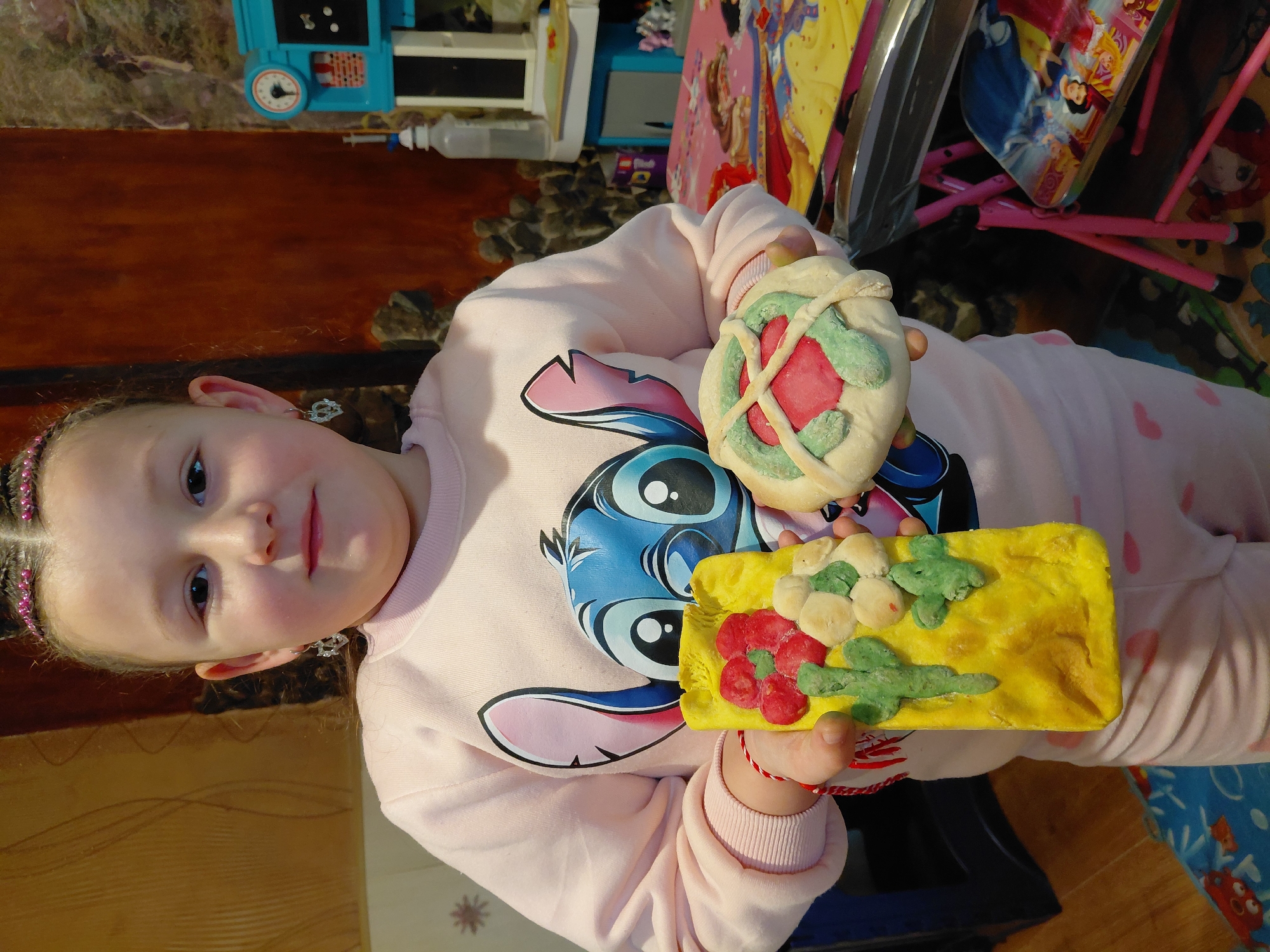 A young girl is holding two pieces of what appears to be artwork crafted from dough or clay, presumably made by her. The girl has fair skin, dark eyes, and her hair is pulled back with colorful braids. She is wearing a light pink sweatshirt with a large cartoon character (Stitch) printed on the front. She also has a red and white bracelet.
In her hands, she holds:
1. A round, bread-like object decorated with red and green shapes, possibly meant to resemble a flower or decorative design.
2. A rectangular, yellow piece with green and red decorations, possibly depicting flowers or animals.
The background is cluttered, with various toys, furniture, and a patterned floor visible. Items like a blue toy kitchen, a Disney-themed item, and pink chairs add to the child-like environment.
There is no information on whether the image was taken in Sofia, Bulgaria. The details are generic and do not indicate this specifically.