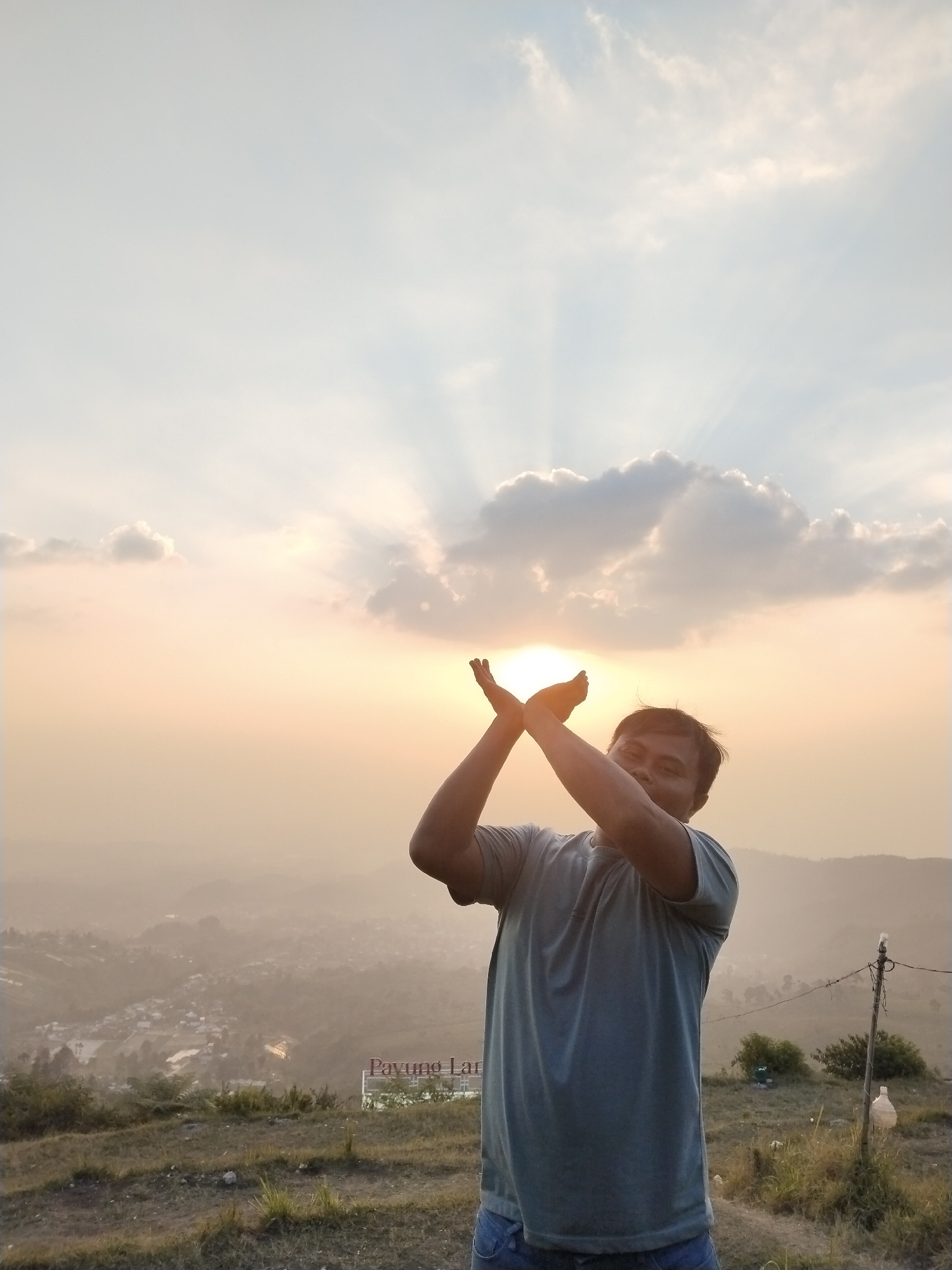 A man stands on a grassy hill overlooking a hazy valley with a town or village below. He is in the foreground, facing the camera and holding his hands up, cupping what appears to be the setting sun between them. The sun is a bright orb, partially obscured by clouds, with sunbeams radiating outwards into the sky. The man is wearing a grey t-shirt and blue jeans. In the background, there is a signpost that reads "Payung Lan". The overall atmosphere is serene and picturesque.