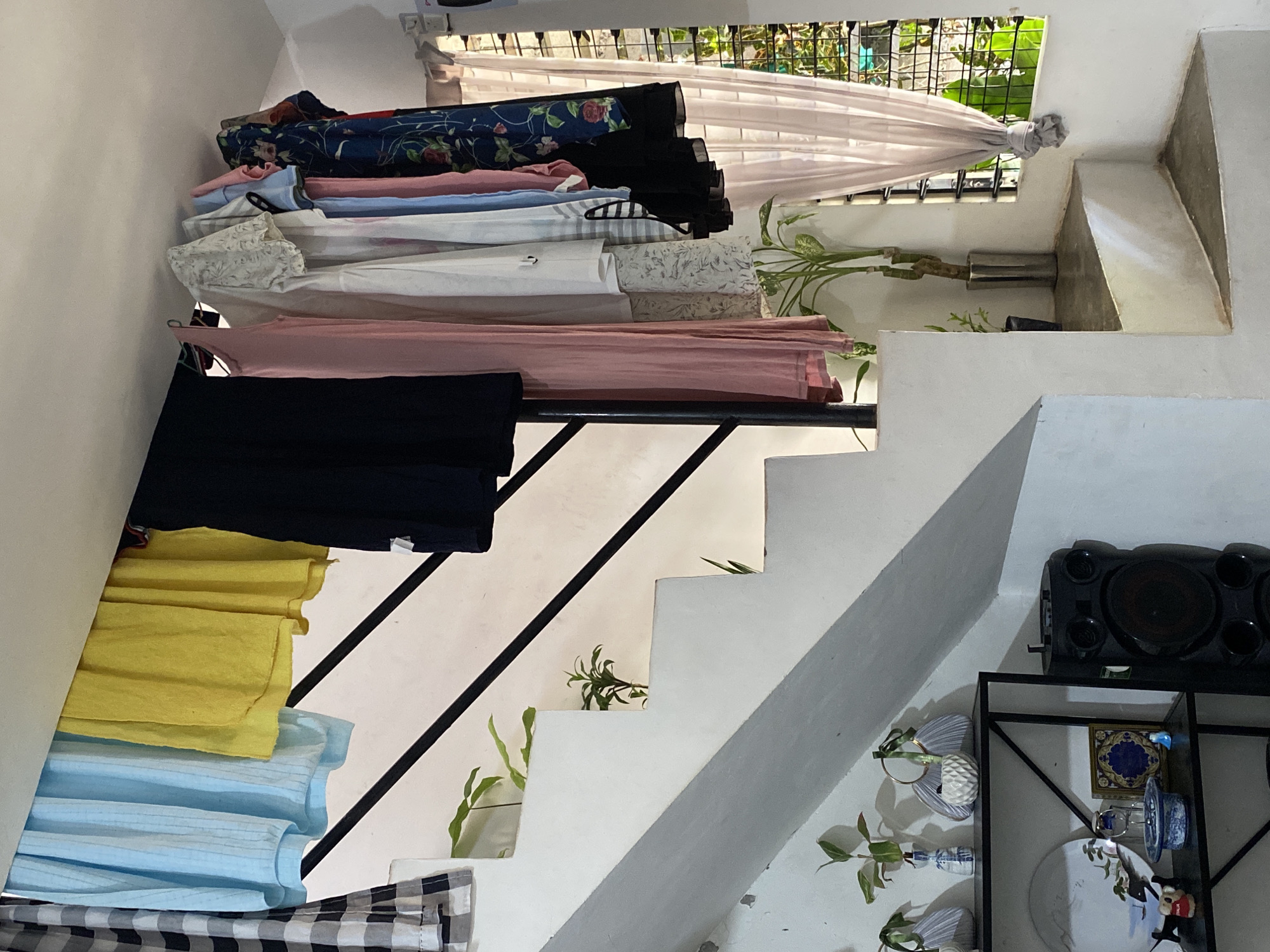 The image shows a multi-level staircase with a metal railing. The staircase is lined with various items, including folded clothing in shades of blue, yellow, pink, white, and black, as well as a patterned fabric. Plants are interspersed along the staircase. A window is visible in the background, covered partially by a light-colored curtain. To the right of the staircase, there is a black shelf unit with a sound system on top, along with decorative plates and other objects. The location is likely an indoor space in Davao City, Philippines, given the tropical vegetation visible through the window.