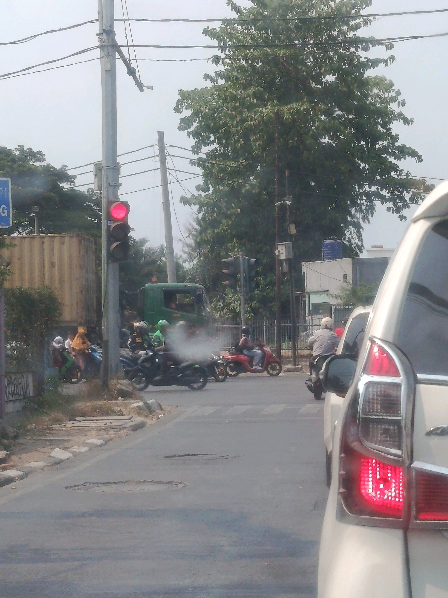 In East Jakarta, Indonesia, a traffic scene unfolds at an intersection. A traffic light displays a red signal, indicating vehicles should stop. Several motorcycles are present, with riders, some wearing helmets and others not. One motorcycle has two passengers, one of whom is holding a child. A large green truck is stopped behind the motorcycles, with a person visible in the driver's seat. In the foreground, the rear of a white vehicle is visible, with its brake lights illuminated. The environment includes trees, electrical poles, overhead wires, a light-colored shipping container, and buildings in the background. The road surface appears to be asphalt and shows signs of wear, including a large pothole. The weather appears overcast.