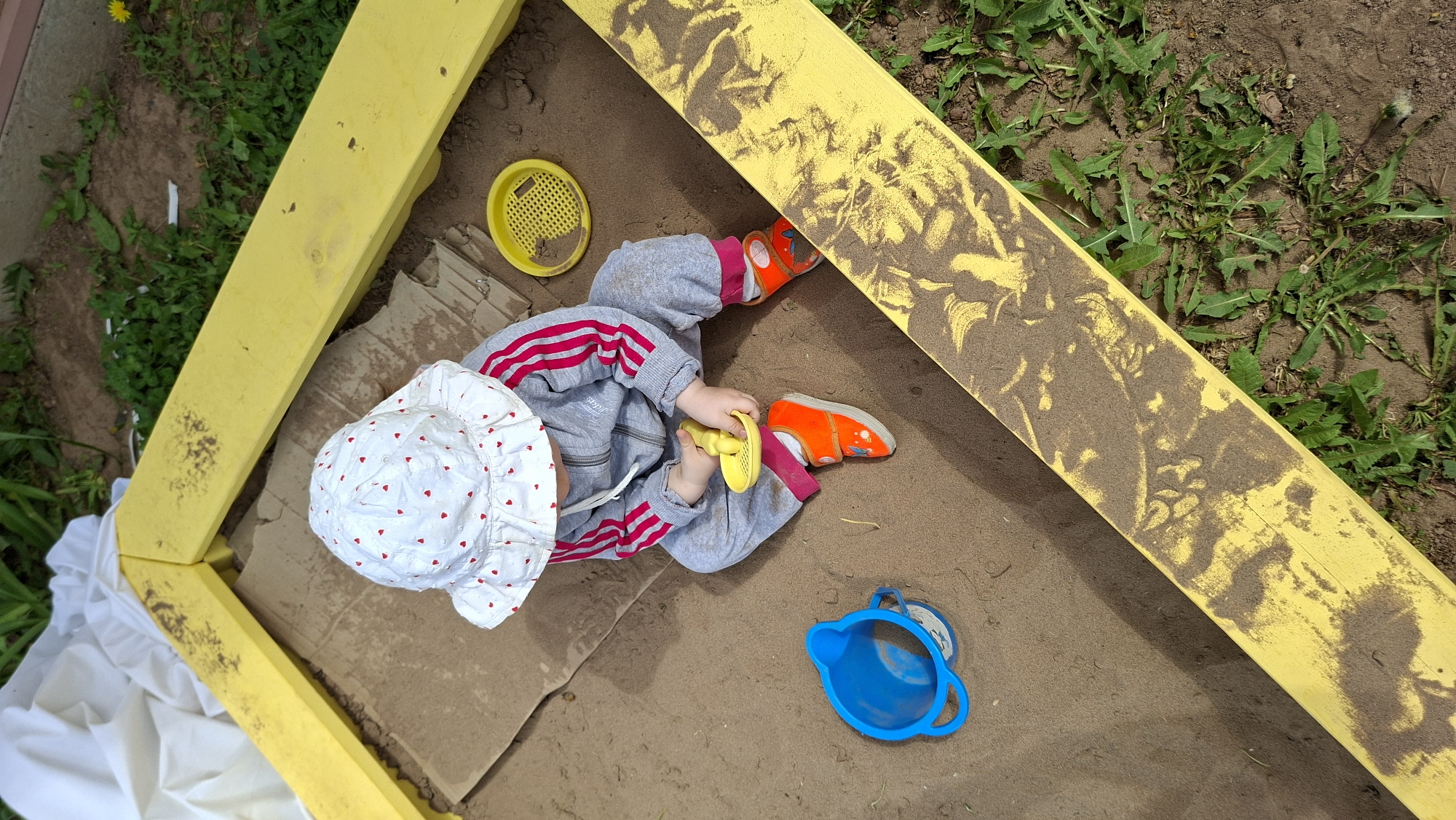 A high-angle, full shot depicts a baby in a sandbox in Большой Букор, Russia. The sandbox has a yellow frame, with traces of sand on the wood. The baby, wearing a white hat with red hearts, a gray tracksuit with red stripes, and orange shoes, sits inside the sandbox. They are holding a yellow toy. A yellow sieve and a blue plastic jug are also in the sandbox. The area surrounding the sandbox has dirt and grass.