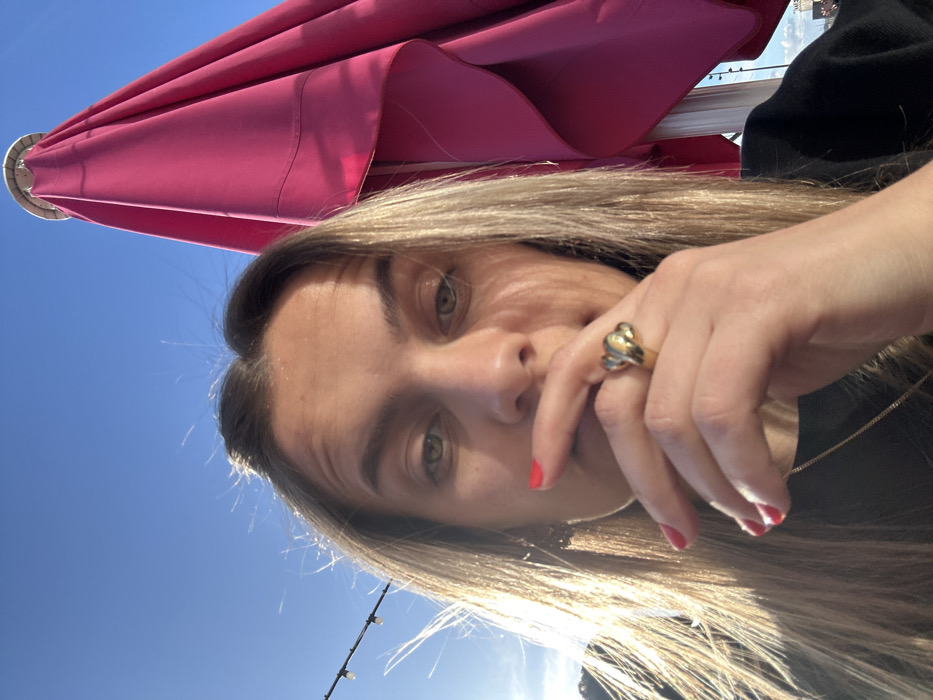 A close-up, slightly upward-angled image, originally rotated 90 degrees counter-clockwise, depicts a person outdoors during daylight. The central subject is an individual with light green or hazel eyes, visible eyebrows, and long, light blonde or brown hair with highlights, appearing somewhat sun-kissed. They are looking directly at the camera with a subtle, neutral to thoughtful expression.
Their left hand is raised, with fingers lightly resting near or partially covering their nose and upper lip. The fingernails are painted with bright red polish. A prominent gold-colored ring, featuring a distinctive braided or intertwined design, is worn on the ring finger of the visible hand. A thin gold chain necklace is visible around their neck, and they are wearing a dark-colored top, likely black or dark blue.
The background on the left side of the frame shows a clear, bright blue sky. A large, vibrant fuchsia pink umbrella, possibly a patio or beach umbrella, occupies the upper right portion of the image. Against the blue sky in the lower left, a string of outdoor decorative lights is visible. The scene is brightly lit by natural sunlight, with the sun appearing to be somewhat behind or to the side of the subject, creating highlights on their hair.