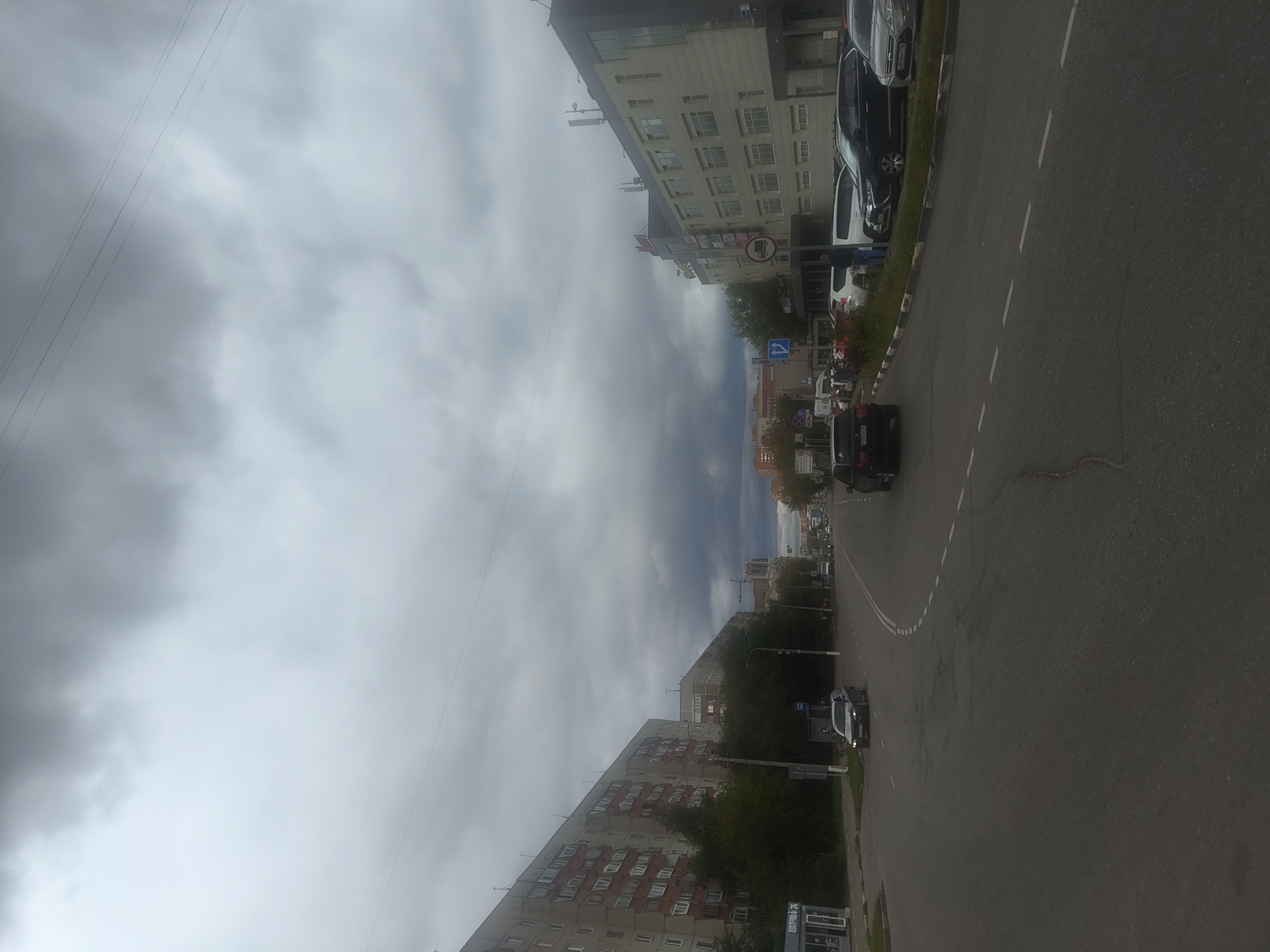 The image depicts a street scene in the city of Zhukovsky, Russia, on a cloudy day. The perspective is from the road, looking down a street lined with multi-story residential buildings on the left and commercial buildings on the right. Several cars are visible, including a dark SUV in the foreground on the right side of the road and a silver sedan further down on the left. The road has multiple lanes and is marked with white lines. Trees and a bus stop are present along the sidewalk on the left. The sky is overcast with heavy, dark clouds.