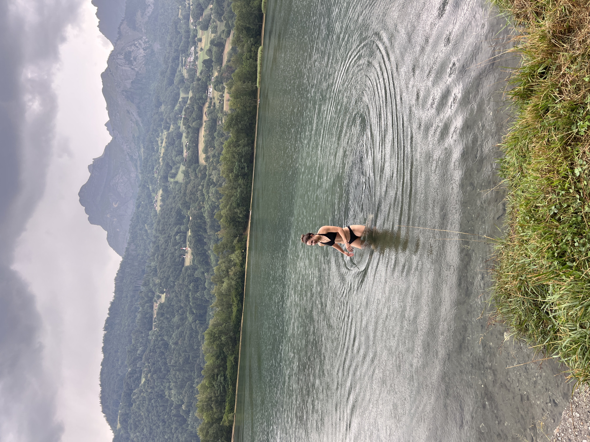 A young woman in a black bikini stands in the shallow water of a lake, creating ripples. She is facing to her right, with her arms slightly bent. The water is a deep green, and the lake extends into the background, lined by a dense forest of green trees. In the distance, a range of mountains rises against a cloudy, grey sky. The foreground shows a grassy bank with some dry reeds and pebbles. The overall scene suggests a natural, outdoor setting, possibly near a recreational area, with the woman enjoying the water despite the overcast weather. The location is the Lake of Passy, situated in the commune of Passy, near the town of Sallanches in the Haute-Savoie department of France.