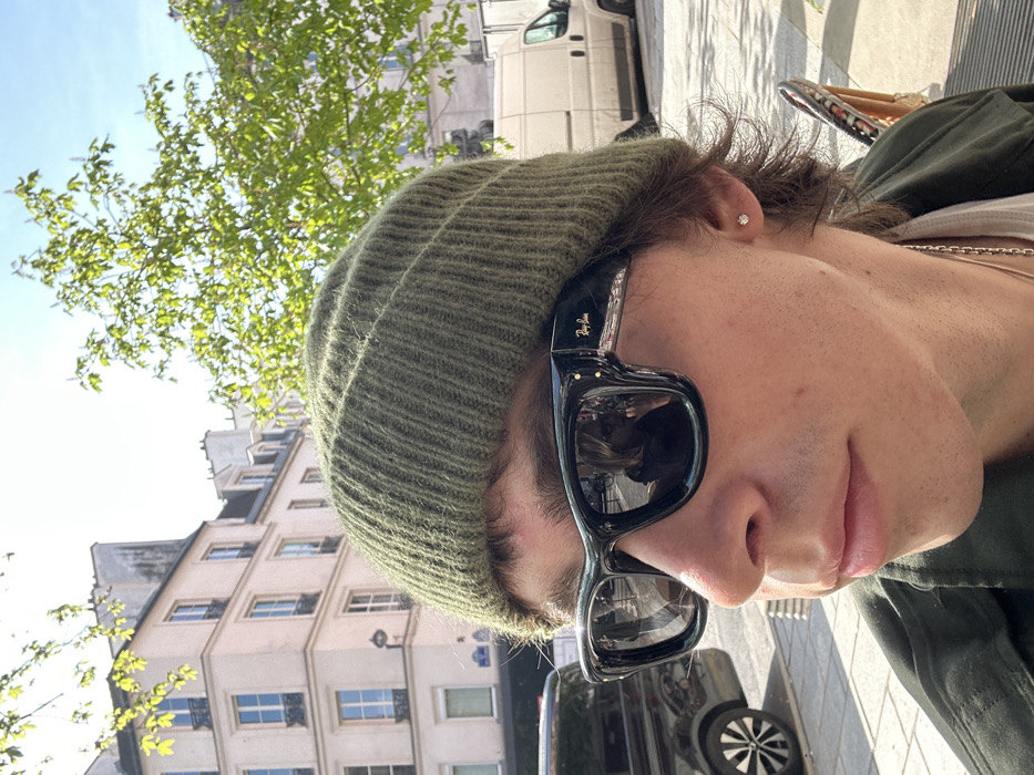The image presents a close-up portrait of an individual situated on a sunny street in Paris, France. The subject wears an olive green ribbed beanie and black, square-framed Ray-Ban sunglasses with visible brand lettering on the right temple. The left sunglass lens reflects a person with blond hair, partially obscured, appearing to hold a device near their face. The subject's face, visible from the nose to the lower jaw, shows fair skin and light stubble. Brown hair is visible extending from under the beanie around the ears, and a small, clear stud earring is present in the left earlobe. The individual is wearing a dark olive green outer garment over a lighter-colored shirt, accented by a silver-toned chain necklace.
The background features typical Parisian Haussmannian architecture, specifically a light-colored, multi-story building with multiple windows, some equipped with black wrought-iron balconies, and a mansard roofline. Bright green foliage from trees is visible, indicative of spring or early summer. On the street level, a dark-colored minivan is partially visible to the left, and a light-colored commercial delivery van is situated further back to the right. Partial blue Parisian street signs are discernable on the building facade. The entire scene is brightly illuminated by direct sunlight, casting clear shadows.