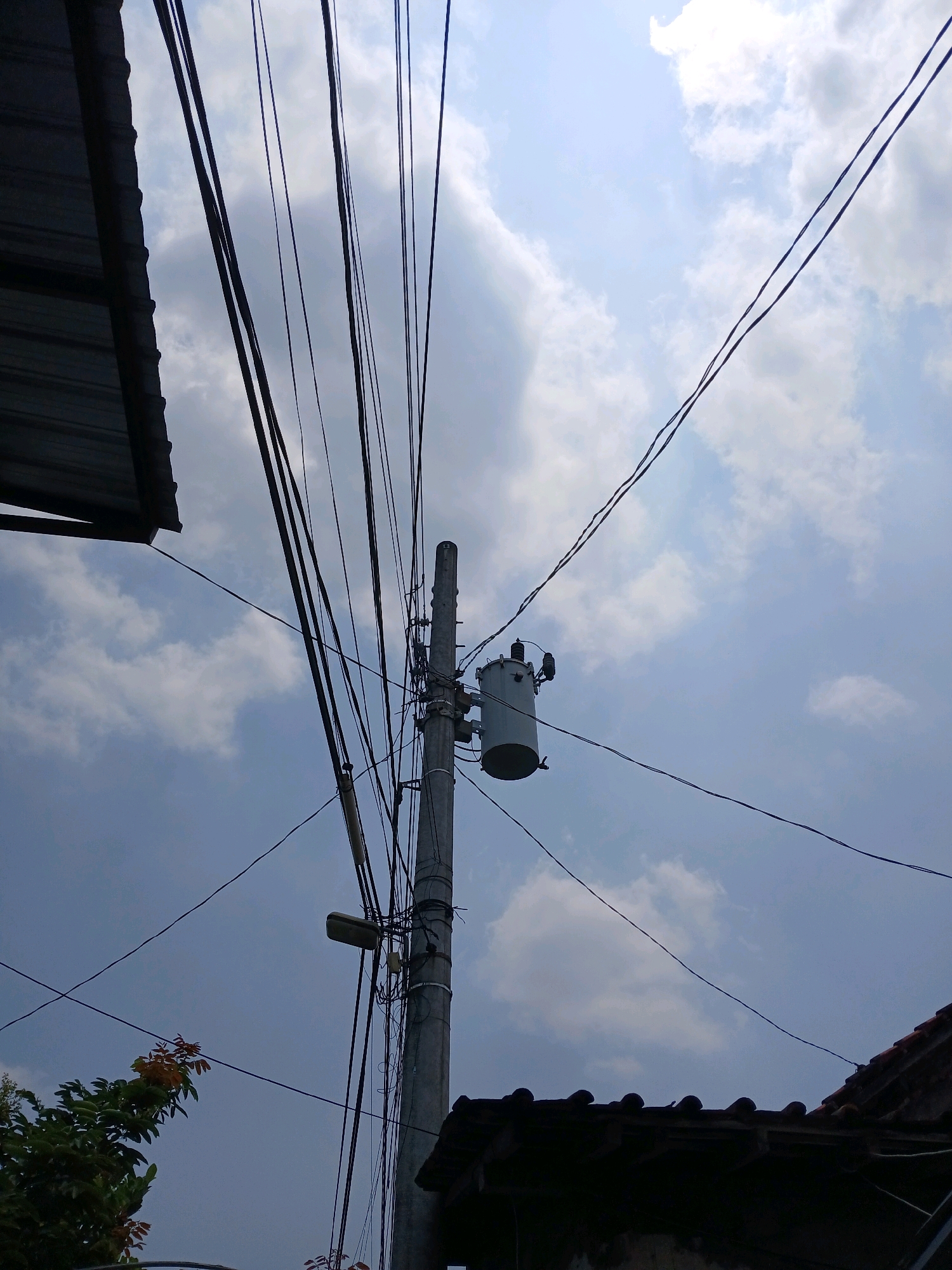This image depicts a utility pole with a transformer and numerous electrical wires against a backdrop of a cloudy sky. The scene is likely located in the city of Yogyakarta, Indonesia, suggested by the presence of typical residential rooftops and vegetation. A metal roof structure is visible on the left, and clay tiled rooftops are present on the right. The utility pole is made of concrete and is cluttered with wires and a street light. A transformer, a cylindrical device with electrical connections, is mounted on the pole. Some foliage from a tree is visible in the lower left corner. The lighting suggests it is daytime. There are no people or specific actions evident in the image.