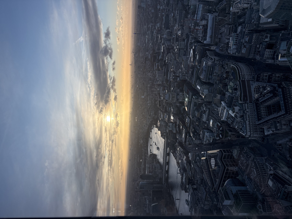 A high-altitude, panoramic view of central London, United Kingdom, captured during sunset. The sun, partially obscured by stratocumulus clouds, is positioned low on the western horizon, casting an intense orange and yellow glow across the sky and cityscape. The River Thames occupies the left-central portion of the frame, flowing east-west, with at least three identifiable bridges spanning it and multiple small vessels visible on its surface. The prominent dome of St Paul's Cathedral is located in the mid-right area of the image. The urban environment below consists of a dense network of buildings, displaying a mix of historical and contemporary architectural styles, extending to the distant horizon under diffused light. The sky transitions from a light blue at the zenith to an orange-yellow gradient near the horizon, interspersed with scattered cloud formations. No individual persons are discernible. A faint vertical dark line along the far left edge suggests the image was captured through a window or structural frame.