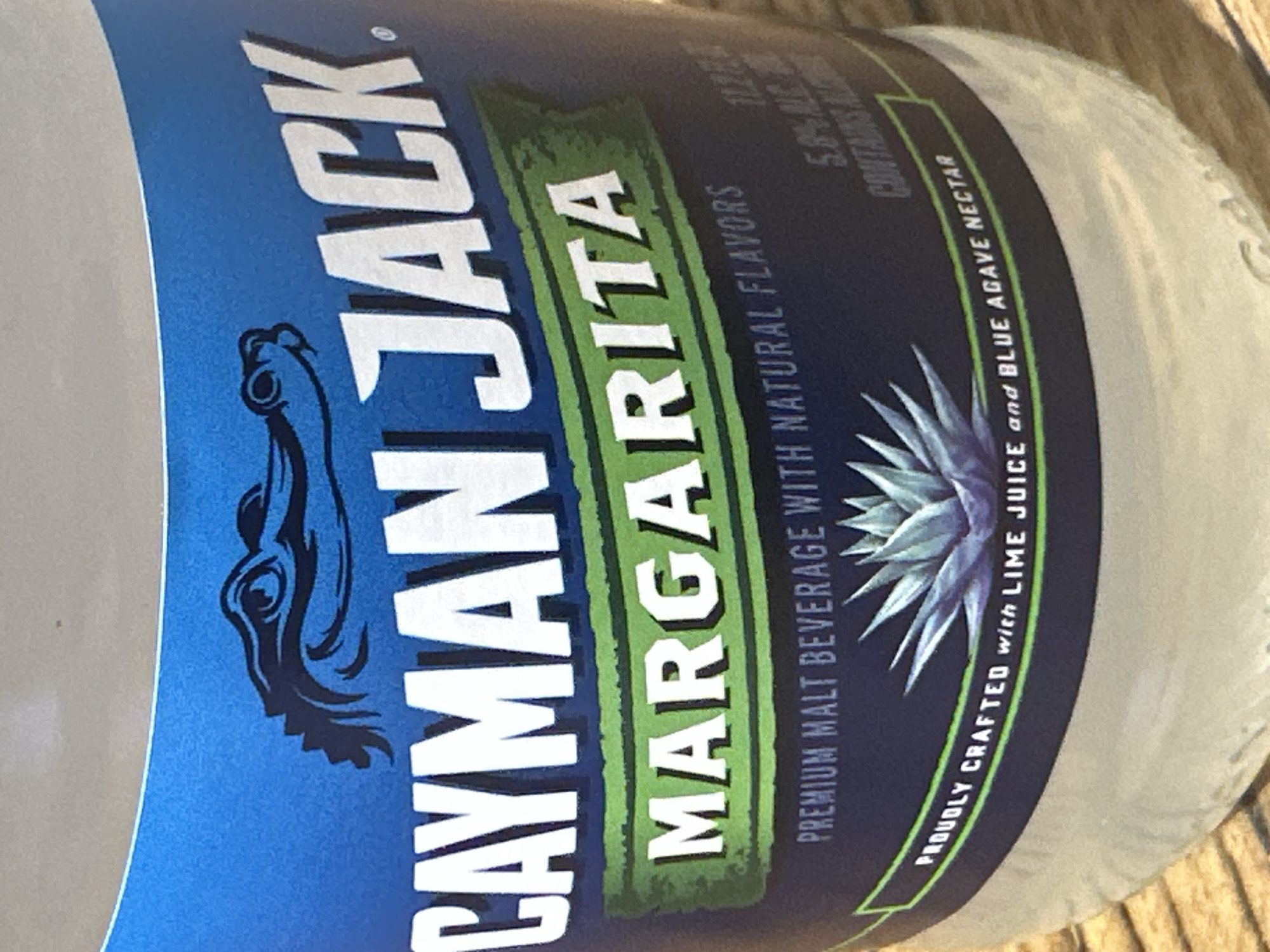 The image is a close-up, slightly angled shot of a glass bottle of Cayman Jack Margarita. The label is predominantly blue and white, with a green accent band. At the top of the label, a black silhouette of an alligator's head is visible. Large white letters spell out "CAYMAN JACK" across the upper part of the label. Below this, on the green band, "MARGARITA" is written in white letters. Further down, smaller text reads "PREMIUM MALT BEVERAGE WITH NATURAL FLAVORS" and "PROUDLY CRAFTED with LIME JUICE and BLUE AGAVE NECTAR." A drawing of an agave plant is located on the lower part of the label. The bottle itself appears to be made of clear glass and has a slight sheen. The background is blurred, but suggests a wooden surface. There is no indication of the city of Camp Hill, United States of America within the image itself.