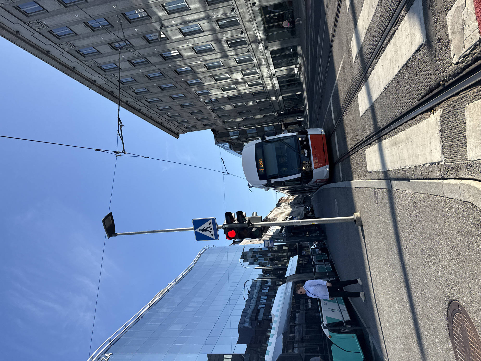A wide shot captures a street scene in Tallinn, Estonia, bathed in sunlight under a clear blue sky. The foreground features a pedestrian crossing with white stripes over tram tracks. To the left, a woman in a light blue collared shirt and black pants stands on the sidewalk, looking towards the left. Adjacent to her is a light blue and white bus with the number "2525" visible. Dominating the center of the frame is a red and white tram approaching the intersection, displaying "4 Lennu" on its destination board. The tram has "CAF" branding. The street is lined with buildings. On the left, a modern glass building reflects the sky and surrounding structures. On the right, a grey, multi-story building with many windows exhibits architectural details. A traffic light with a red signal is visible on a pole next to a pedestrian crossing sign. Overhead, power lines for the tram run across the sky.