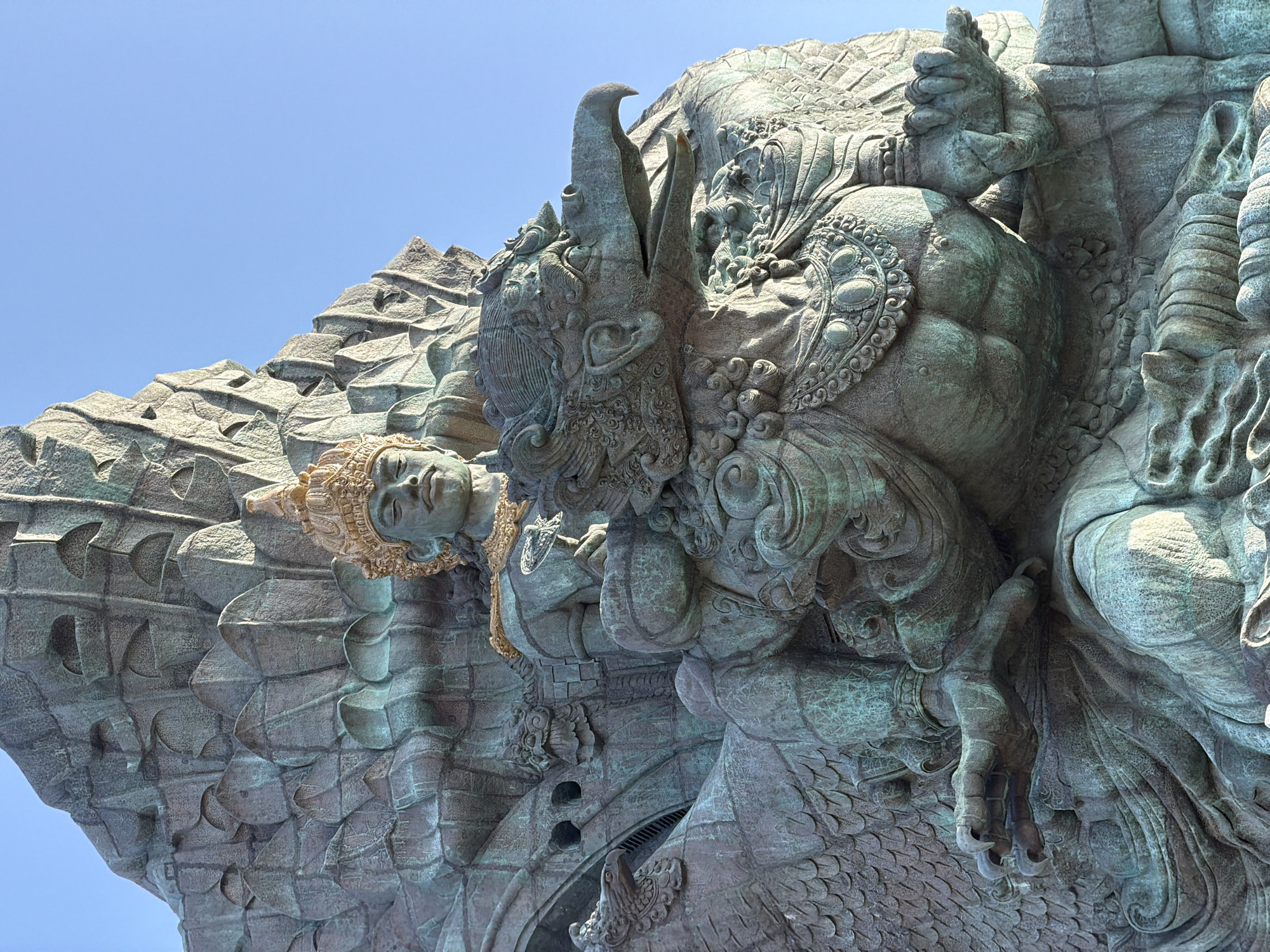 The image shows a close-up, low-angle view of the Garuda Wisnu Kencana statue in Kuta Selatan, Bali, Indonesia. The statue is made of a dark, weathered material, likely bronze or stone, with a greenish patina. It depicts a human figure with a golden headdress and ornate jewelry, seated upon a large, eagle-like creature, presumably Garuda. The Garuda has a detailed beak, feathered wings, and talons. The background is a clear, bright blue sky, indicating daytime. The statue is massive and intricately carved, with details visible in the facial features, clothing, and feathers. The lighting suggests the sun is high, casting shadows that highlight the sculpture's textures and form.