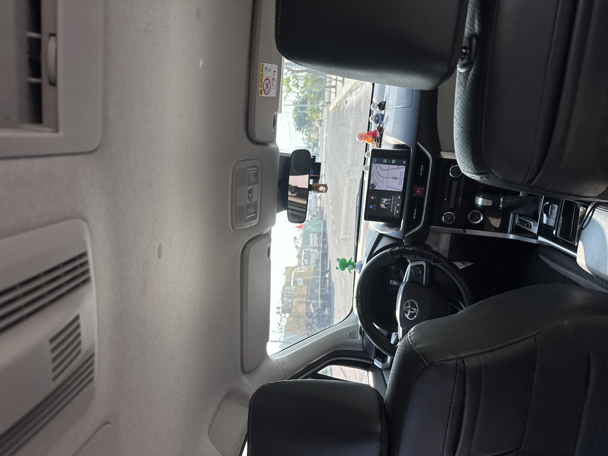 The interior of a car, likely a Toyota, is shown from an elevated perspective, focusing on the driver's area. The dashboard is equipped with a touchscreen navigation system displaying a map. The steering wheel, adorned with the Toyota logo, has a green frog toy hanging nearby. The gear shift and central console are visible, along with the front seats upholstered in black leather. The rearview mirror hangs from the windshield, and the sun visor is positioned above. Outside, a street scene is visible through the windshield, suggesting a location in an urban area. There is a building with a yellow body.