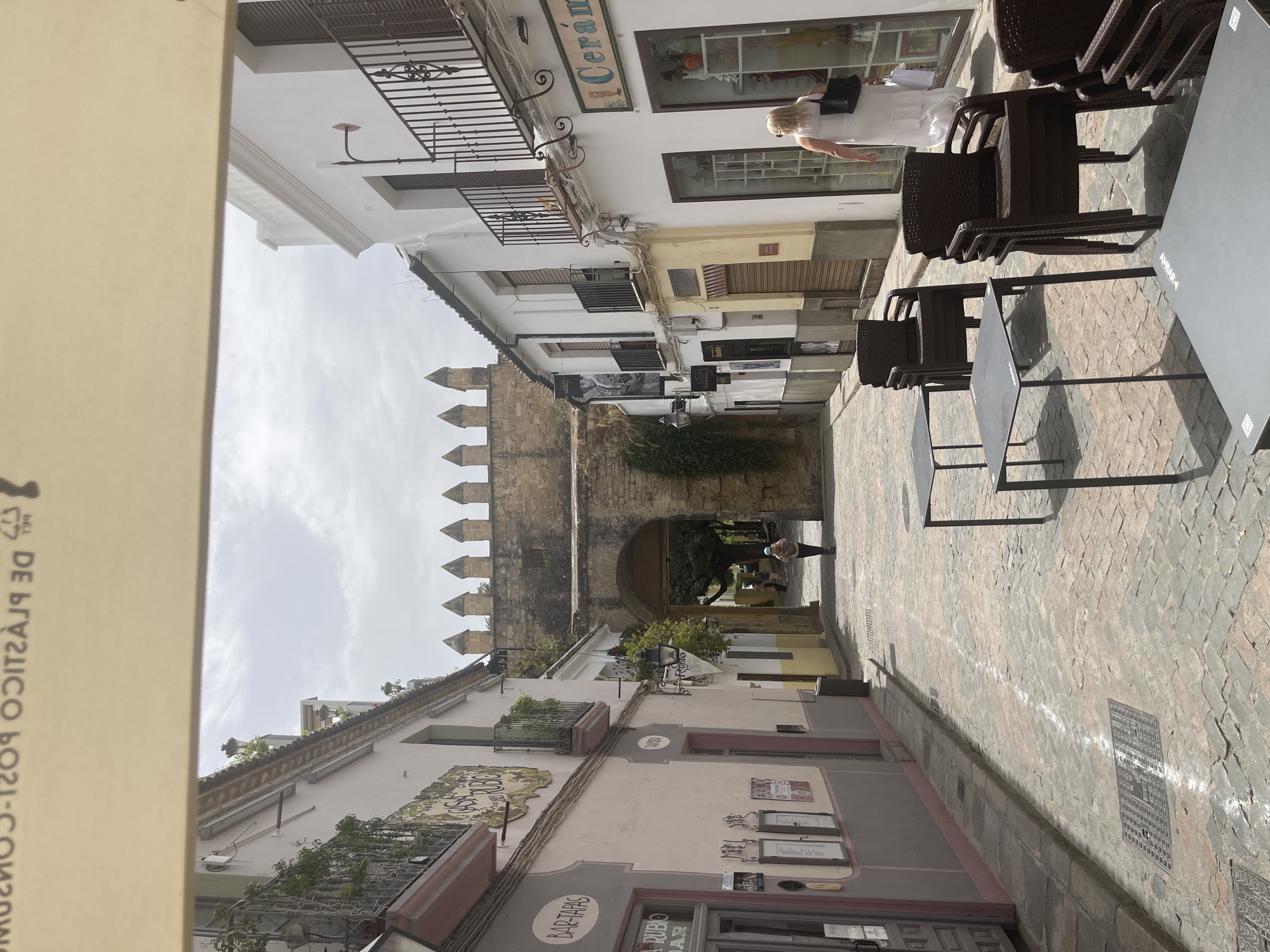 This media file depicts a view of a narrow, cobblestone street in Córdoba, Spain, likely during daylight hours. The scene is characterized by historic architecture, with buildings lining both sides of the street, featuring traditional balconies and ornate details. To the left, a light-colored awning partially obscures a building, with some text visible on its surface. The buildings display a mix of architectural styles, some appearing older with stone elements, while others are more modern with white facades and dark-framed windows.
In the distance, an arched gateway, part of an ancient wall with crenellated battlements, suggests a historical significance to the area, possibly leading into a courtyard or another part of the old city. On the right side of the street, several dark brown, stackable chairs and dark tables are set up, indicating an outdoor seating area for a cafe or restaurant. A dark-colored trash receptacle is also visible.
There are a few individuals present. Near the top right, a woman with blonde hair, wearing a white dress and carrying a dark bag, is seen walking past a shop window. Further down the street, near the arched gateway, a person is visible from behind, appearing to be walking away.
The weather appears to be partly cloudy, with visible clouds in the sky. The overall atmosphere seems relaxed and typical of a European city on a pleasant day, with the cobblestone streets and historic buildings contributing to a sense of timelessness.
Several pieces of text are visible, though some are partially obscured or difficult to read. On one building, the word "Cerámica" is prominently displayed on a sign. Another sign reads "BAR-TAPAS." Other signs on storefronts and menus are present but not clearly legible. The text on the awning on the left is in Spanish and appears to be a sustainability-related message.