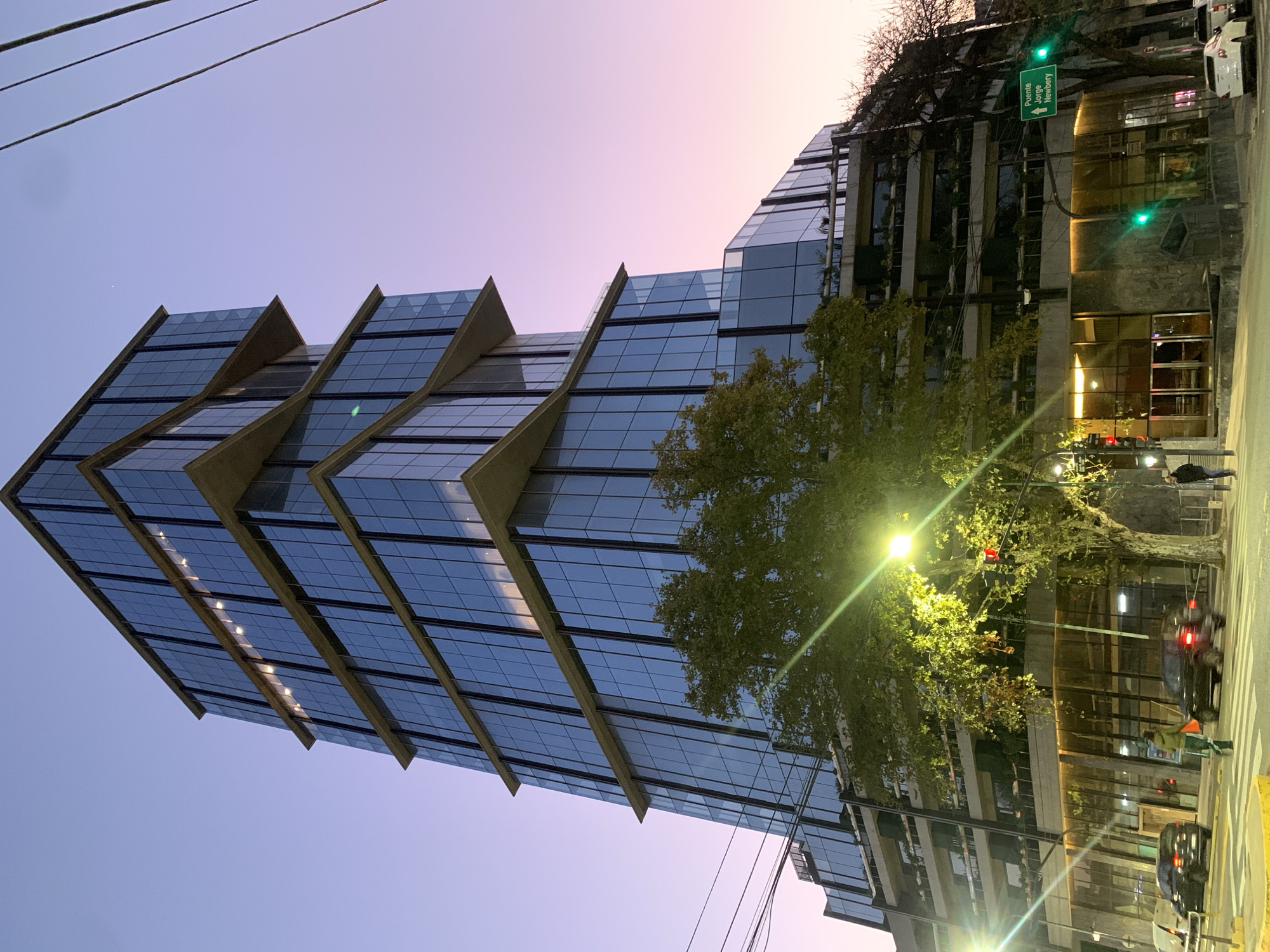 The image presents a nighttime scene of a modern, high-rise building located on a street in Buenos Aires, Argentina. The building is characterized by a staggered design, featuring reflective glass windows and concrete balconies that jut out at each level. The sky transitions from a light blue to a faint pink at the top of the frame, suggesting either dusk or dawn. A street sign visible in the upper right indicates directions to "Puente Jorge Newbery."
On the street level, a tree with illuminated leaves and a street lamp are prominent. Pedestrians are walking on the sidewalk near the building, and cars are visible on the street. The general atmosphere is urban, with the modern architecture of the building contrasting against the more natural elements of trees and streetlights. Power lines are also visible in the upper left corner.