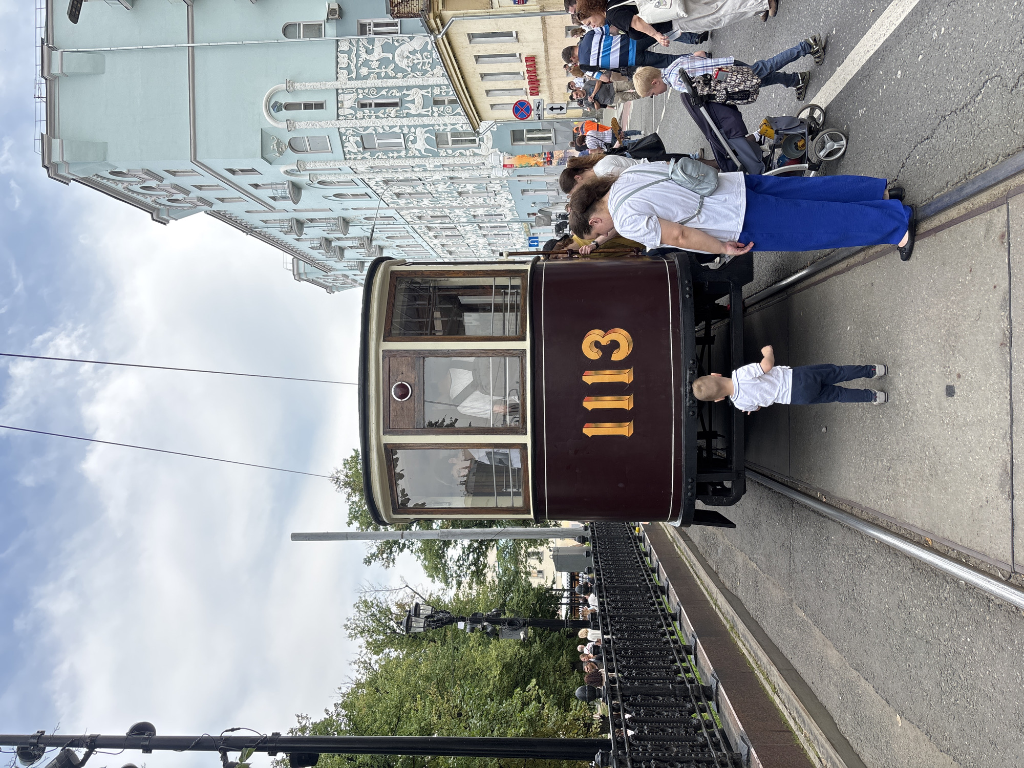 The image depicts a vintage tram, numbered "1113", on a city street in Moscow, Russia. The tram is a deep maroon color with gold numbering and appears to be on display or part of an event, as a crowd of people are gathered around it. In the foreground, two young boys are looking at the tram, one from the rear and another from the side. A woman in blue pants and a white shirt is standing near the tram, facing it. Behind her, a person is pushing a stroller with a child in it. A man in a striped shirt is visible in the background, walking away from the tram. The street is paved with asphalt and concrete, with tram tracks running down the center. To the left of the tram, a black ornate metal fence runs along a raised embankment covered with grass and trees. In the background, a light blue building with intricate white decorative elements and arched windows dominates the cityscape. Several other buildings, including a yellow one with a red sign, are also visible. The sky is overcast with scattered clouds. The scene suggests a public gathering or historical reenactment centered around the vintage tram.