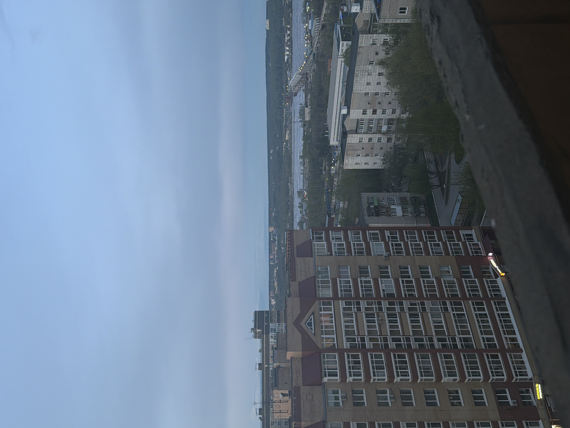 The image captures a high-angle view overlooking Perm, Russia, likely taken from a tall building or structure. The sky is predominantly blue, suggesting dawn or dusk. A river stretches across the middle ground, bordered by dense trees on the near bank and buildings on the opposite bank. A bridge spans the river, illuminated by lights. Several tall buildings are visible, including a large brick building with many windows. The overall scene suggests an urban environment. The perspective indicates the viewer is elevated, looking out over the city.