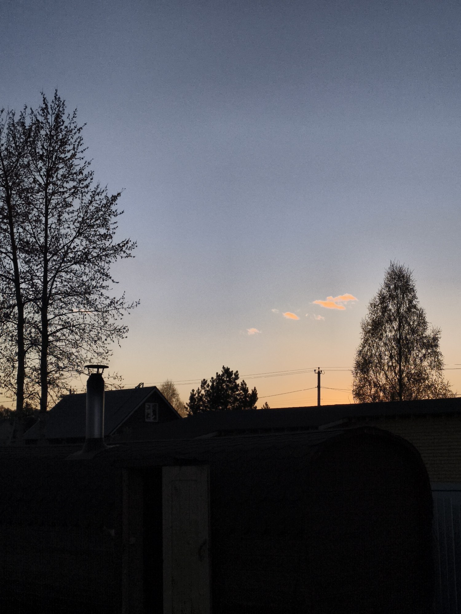 The image depicts an outdoor scene in Русский Вожой, Russia, at twilight. The sky transitions from a pale blue at the top to orange near the horizon, with a few scattered clouds illuminated by the setting sun. Silhouetted trees are visible on the left and right sides. In the foreground, a dark, rounded structure obscures much of the view. Behind this structure, there is a building with a visible roof and a chimney. Power lines and a utility pole are also present in the background. The overall setting suggests a rural or suburban environment.
