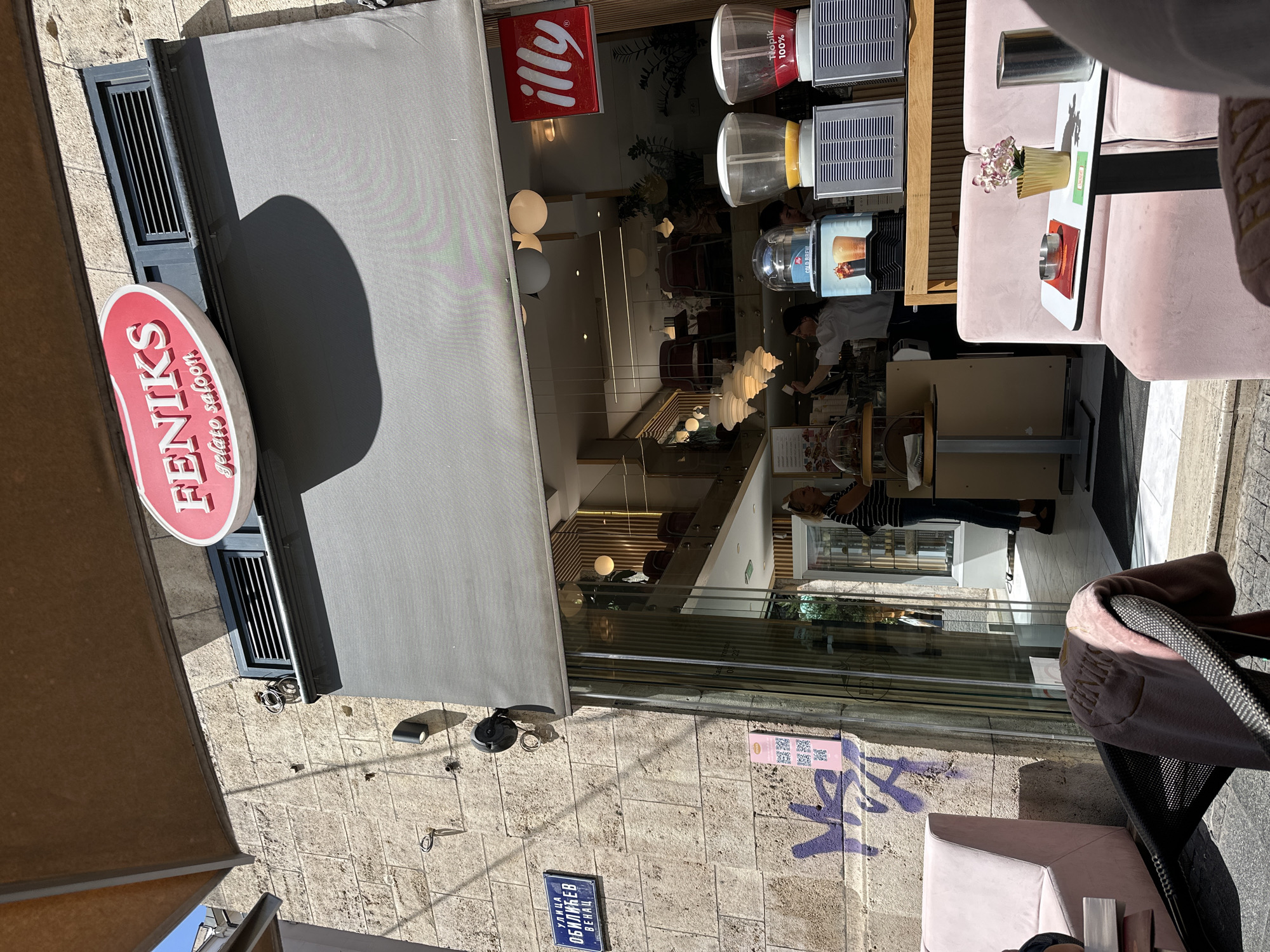 The image shows the exterior and partial interior of "Feniks Gelato Salon" in Belgrade, Serbia. A red and white sign for Feniks is visible. The establishment has a gray awning. Inside, a person wearing a striped shirt is visible behind the counter. There are tables and chairs inside the cafe. The Illy coffee logo is displayed near the entrance. On the sidewalk, there are pink chairs and tables, as well as a chair covered with a pink blanket. Graffiti is present on the wall below the store window, and a blue street sign with Cyrillic text is on the lower left. The image appears to be taken during daylight hours.