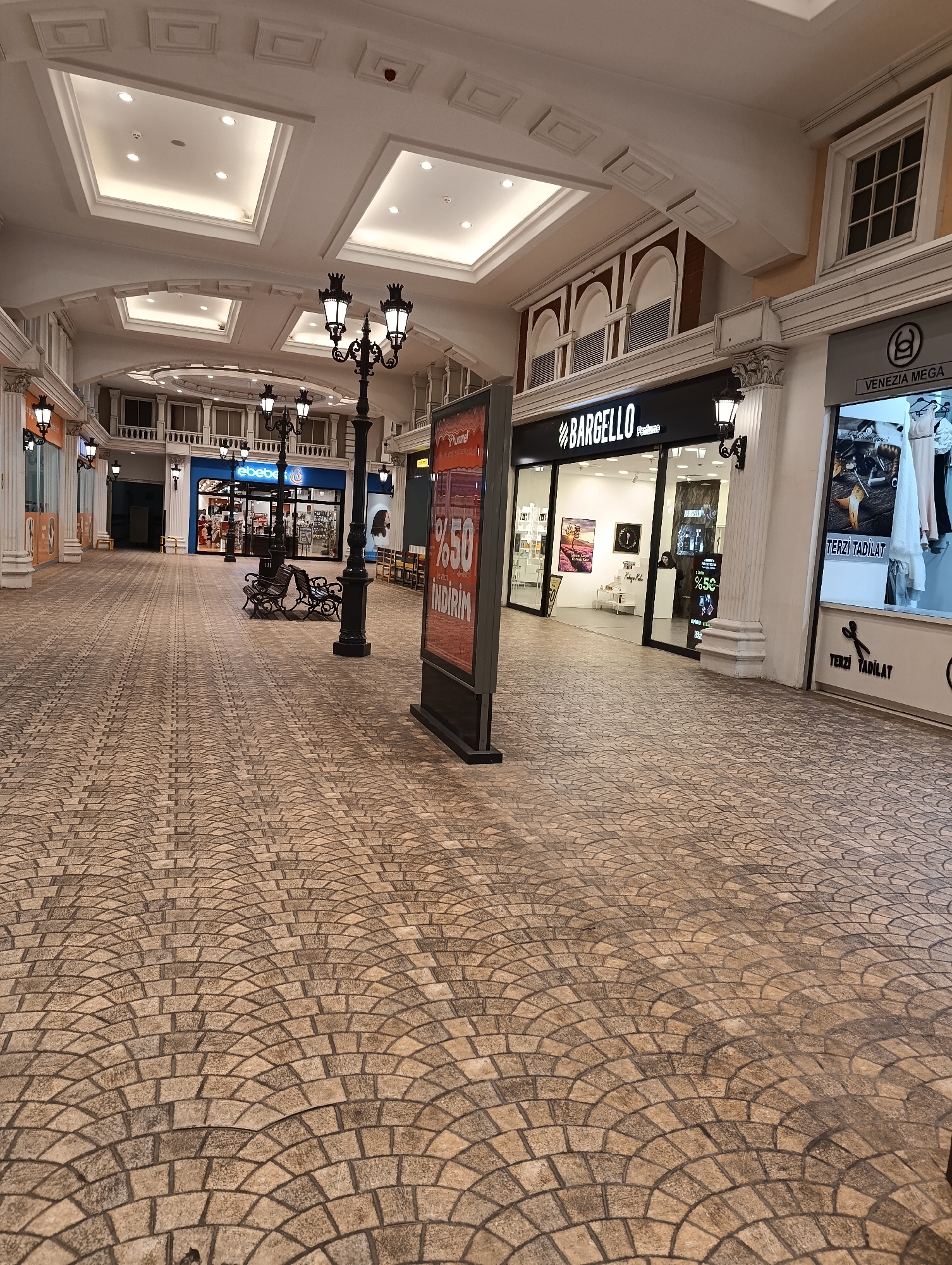 The image shows a section of an indoor shopping mall with a paved floor arranged in a fan pattern. The mall features ornate architecture with white columns, arched ceilings with recessed lighting, and decorative street lamps. Several storefronts are visible, including "BARGELLO Perfume" and "VENEZIA MEGA". One storefront has a sign indicating "50% INDIRIM" (50% discount) and "TERZI TADILAT" (Tailor alteration). A display board with similar discount information is positioned in the foreground. Benches are placed along the walkway. The environment is well-lit and appears to be a commercial area. This scene is located in the Venezia Mega Outlet shopping mall in Gaziosmanpaşa, Istanbul, Turkey. There are no people clearly visible in the image.