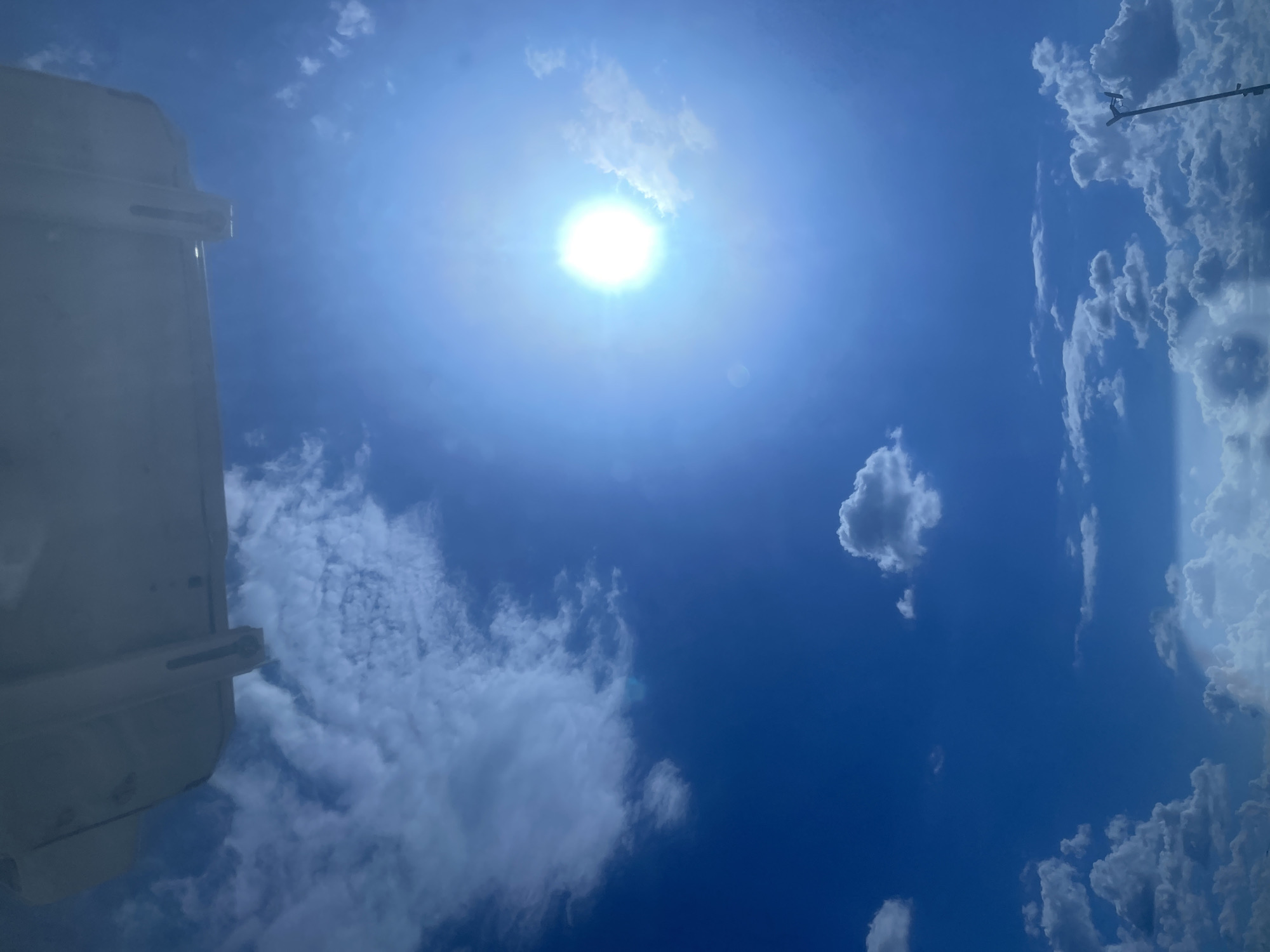 The image depicts a bright, sunny sky with scattered white clouds. The sun is prominently visible in the upper right quadrant, emitting a strong glare. A portion of a white, rectangular structure, possibly the underside of an aircraft or a building overhang, occupies the top portion of the frame. The lower part of the image shows a landscape of clouds, with a faint hint of a rainbow visible in the bottom left corner. A tall, thin object with two lights on top, resembling a street lamp or a fixture, stands on the right side of the frame. The scene appears to be an outdoor environment. There is no information in the image to indicate the location is Podkletnoe, Russia.