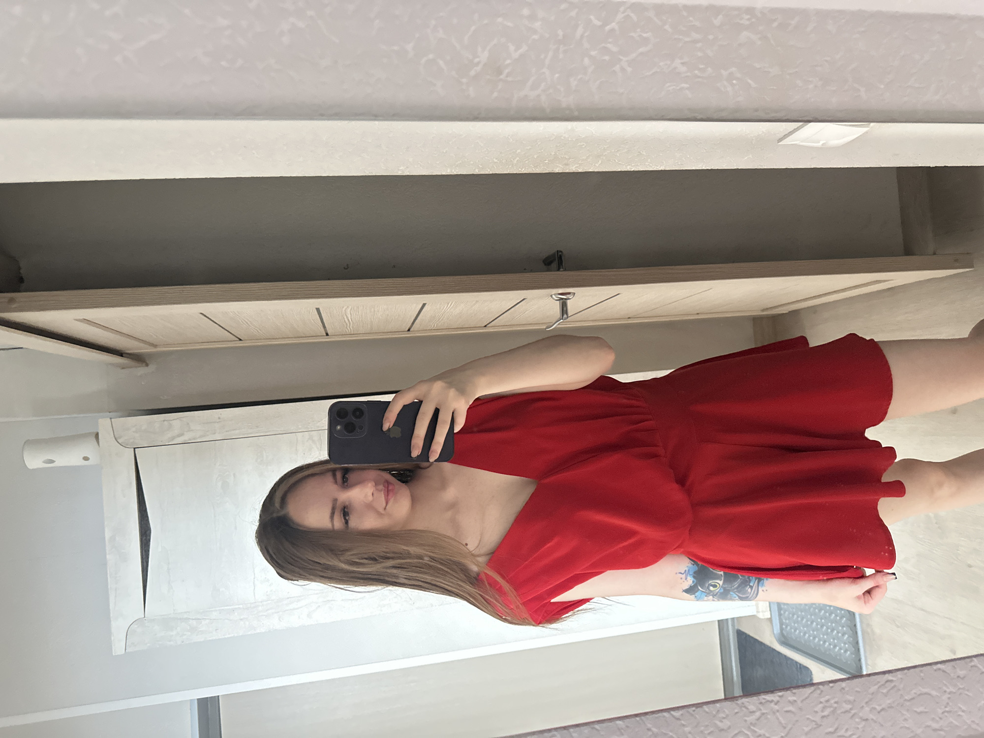 A young woman with long, brown hair and fair skin stands in front of a mirror, taking a selfie with her phone. She is wearing a vibrant red V-neck dress with short, puffed sleeves. The dress has a cinched waist and flares out into a short skirt. She has a tattoo of a black cat with blue accents on her left forearm.
The background shows a reflection of a room with wooden flooring and a light-colored door with horizontal slats. To the left, a tall white wardrobe with black accents is partially visible. The walls appear to be painted white, with some textured wallpaper on the right side of the frame. The overall lighting suggests an indoor setting, possibly a bedroom or dressing area.