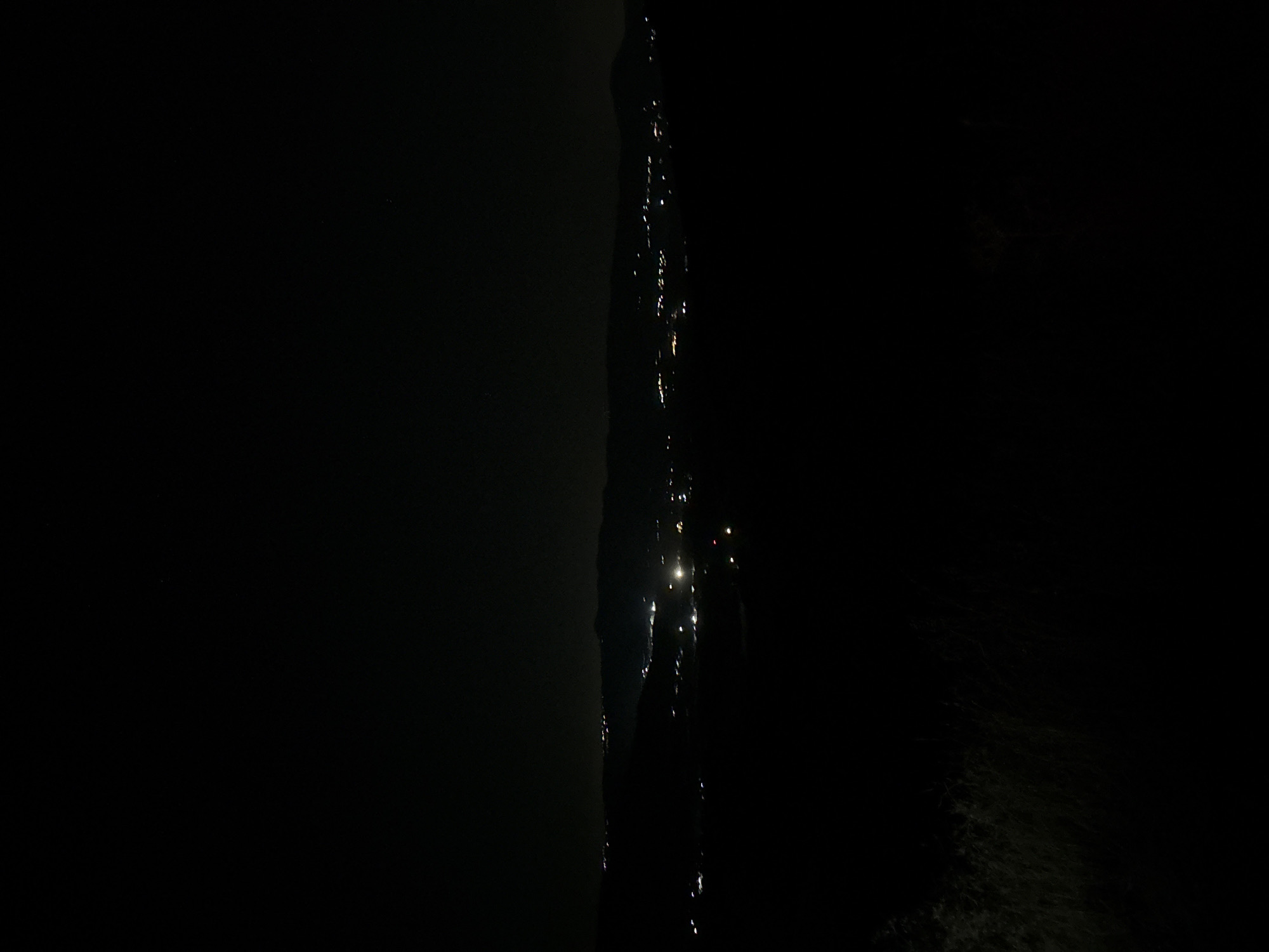 The image is a low-light, wide-angle shot of a landscape at night. The foreground is dark, with some blurry vegetation visible at the bottom left. In the middle ground, a distant settlement is illuminated by numerous scattered lights, suggesting a town or city spread across rolling hills or mountains. The lights vary in intensity and color, with some appearing bright white and others more yellow or orange, indicating different types of lighting sources. The background consists of dark hills or mountains silhouetted against the night sky, which is uniformly dark with no visible stars or moon. The overall impression is of a sprawling urban area at a distance, seen from a vantage point on a dark hillside. The location is the Armenochori Community, Cyprus.