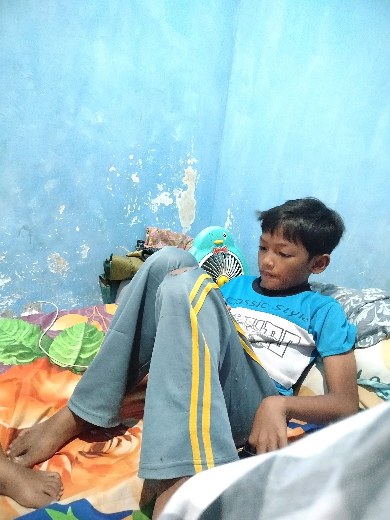 A young boy sits on a bed in a room with a blue, textured wall. He is wearing a blue t-shirt with a graphic print and gray pants with yellow stripes. His legs are crossed, and he is looking downwards. In the background, there is a teal-colored fan shaped like a penguin and some folded clothes. The bed is covered with colorful patterned blankets and pillows. A white electrical cord is visible on the left side of the bed. The scene suggests a casual indoor setting, possibly a bedroom in a residential area of Beloh, Indonesia.