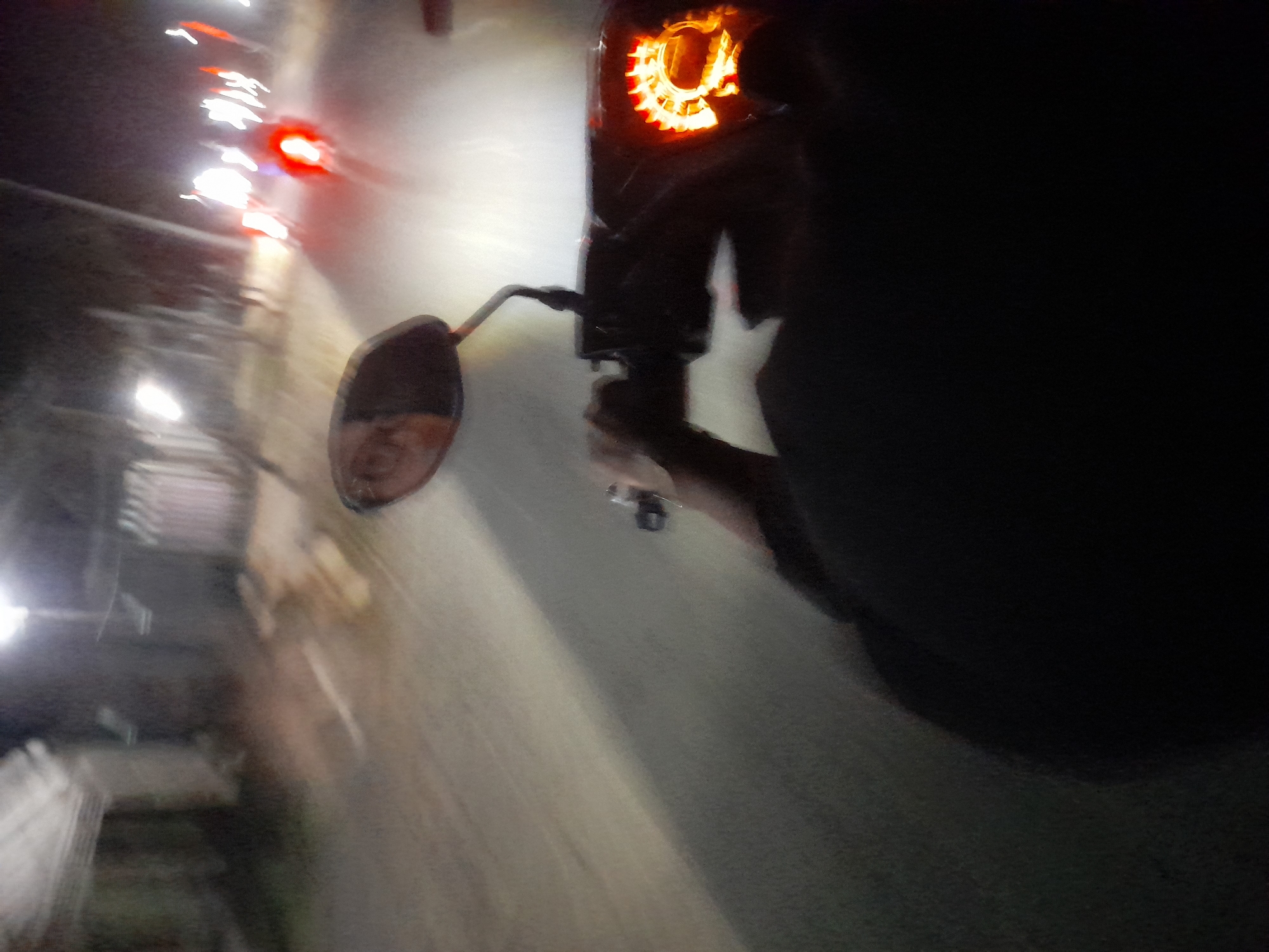 The image depicts a first-person perspective of someone riding a motorcycle at night on a paved road in Jetaksari, Indonesia. The road appears to be illuminated by streetlights and the headlights of other vehicles. The rider's hands are visible on the handlebars, and a rearview mirror is mounted on the left side of the handlebars. The motorcycle's dashboard is lit with an orange glow. In the background, other motorcycles and a building are visible, all blurred due to motion. The scene suggests a typical night scene on a road in Indonesia, possibly in a semi-urban or rural area.
