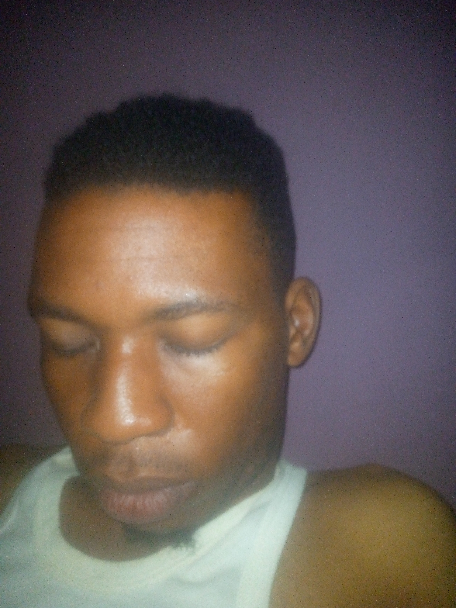 An eye-level close-up captures a dark-skinned Black male with closed eyes and a close-cropped haircut. He is wearing a white tank top. The background is a solid, blurry shade of lavender. There are no elements in the image to indicate the location is Ikorodu, Nigeria.