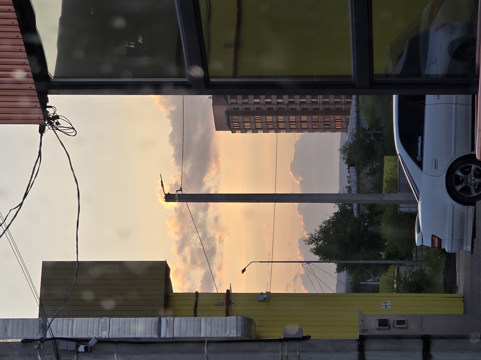 The image, captured in Chita, Russia, shows an urban scene at sunset. The sky is filled with warm hues of orange and yellow, with scattered clouds illuminated by the setting sun. In the foreground, there are elements of urban infrastructure, including what appears to be a corrugated metal roof on the left and a yellow structure on the right, both reflecting the sky's colors. Wires and a utility pole are also visible, suggesting the presence of electrical infrastructure.
In the middle ground, a tall, multi-story building with many lit windows stands against the sunset, hinting at a residential or office building. To the right, a white car is parked, its reflection visible in a mirrored surface above, possibly a window or a display. The car has a sticker on its rear window that reads "@aimettilazta".
The overall impression is one of a typical evening in a city, with the natural beauty of the sunset contrasting with the man-made structures. The lighting suggests it is late afternoon or early evening. There are no people clearly visible in the image. The weather appears to be clear apart from the clouds.