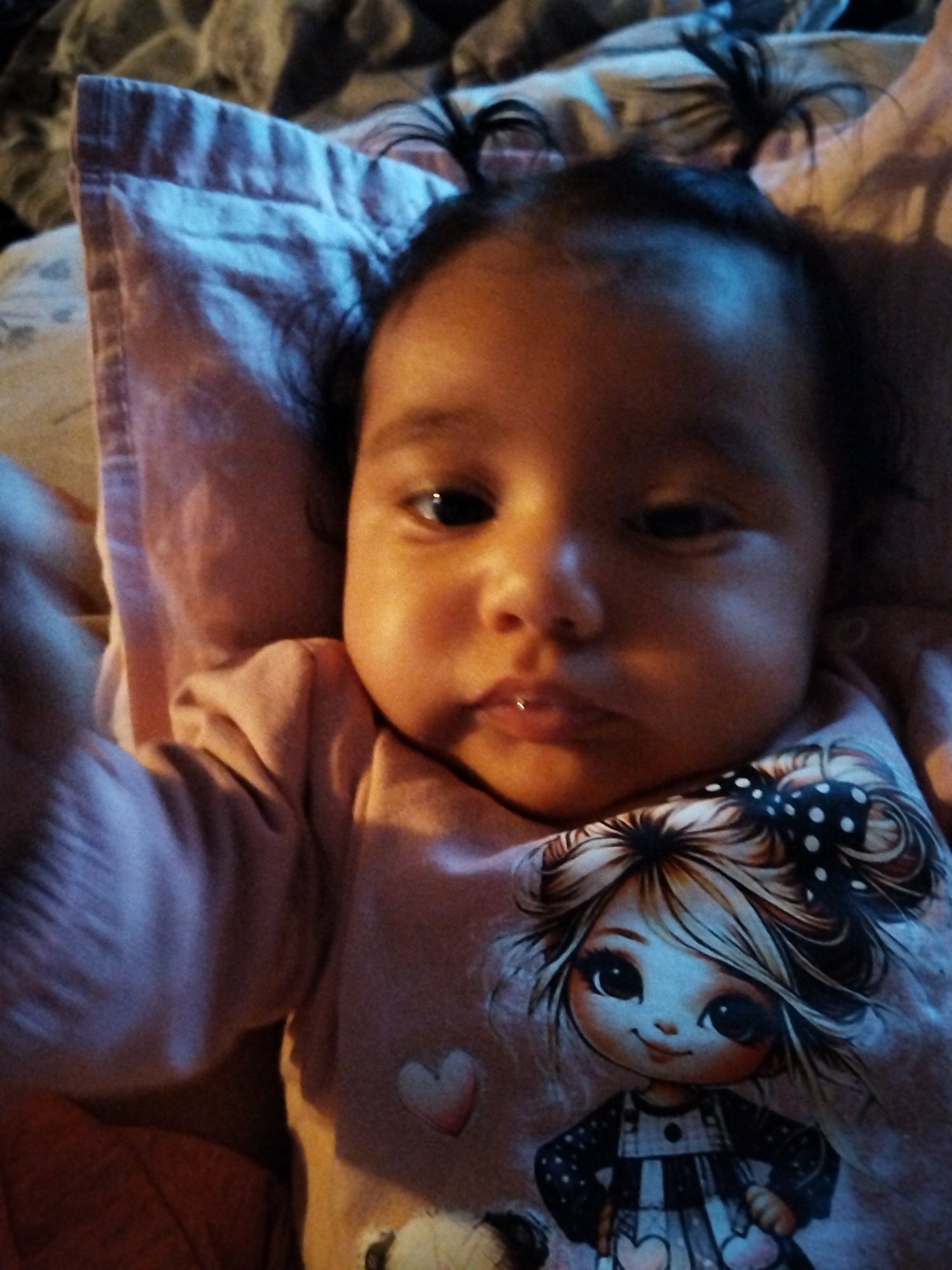 A baby with dark hair in two small ponytails is lying on a light pink pillow. The baby is wearing a light pink long-sleeved shirt with a cartoon character printed on it. The baby is looking at the camera. The background is blurry and appears to be bedding. Location: Unknown, Unknown.